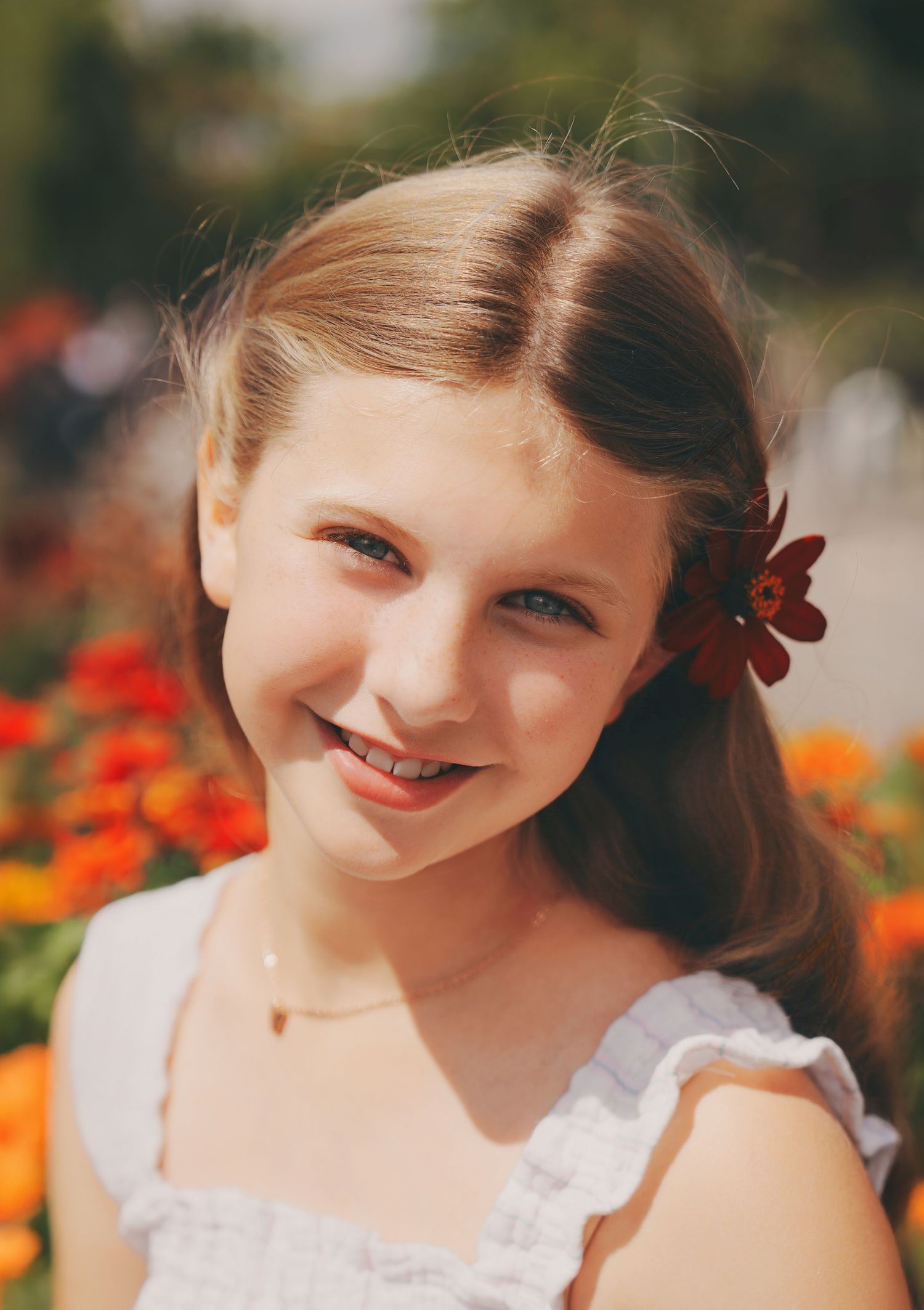 Smiling young girl with red flower in hair poses among colorful blooms in white dress.