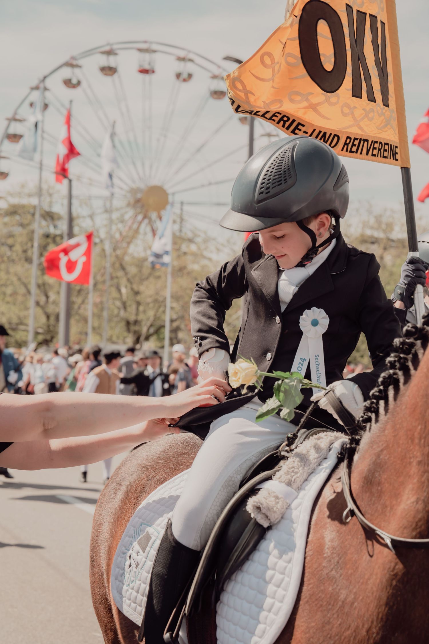 Young equestrian in black riding attire sits on horseback during a parade with a Ferris wheel backdrop.