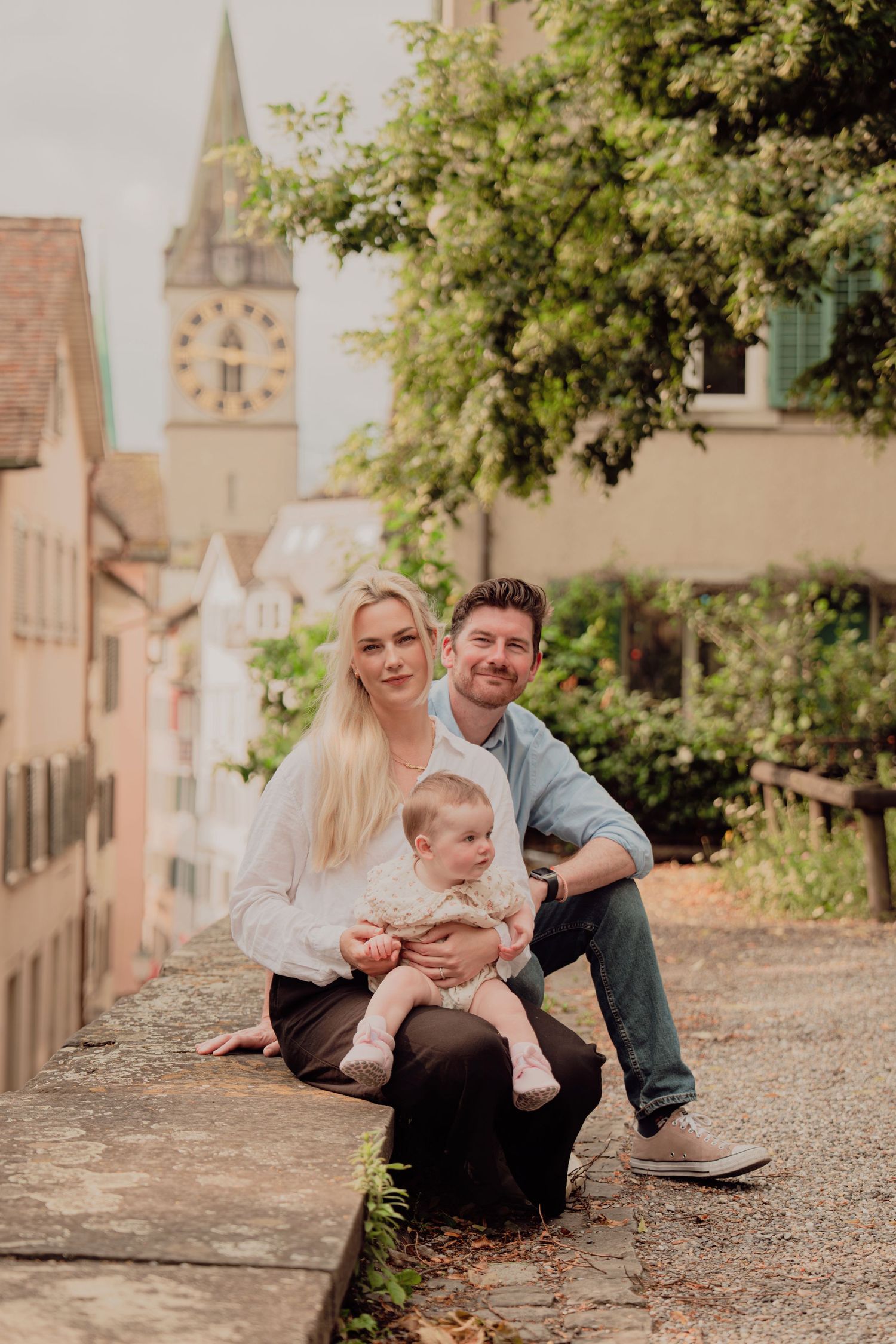 Young family with baby posing on stone steps in a charming European old town with clock tower.