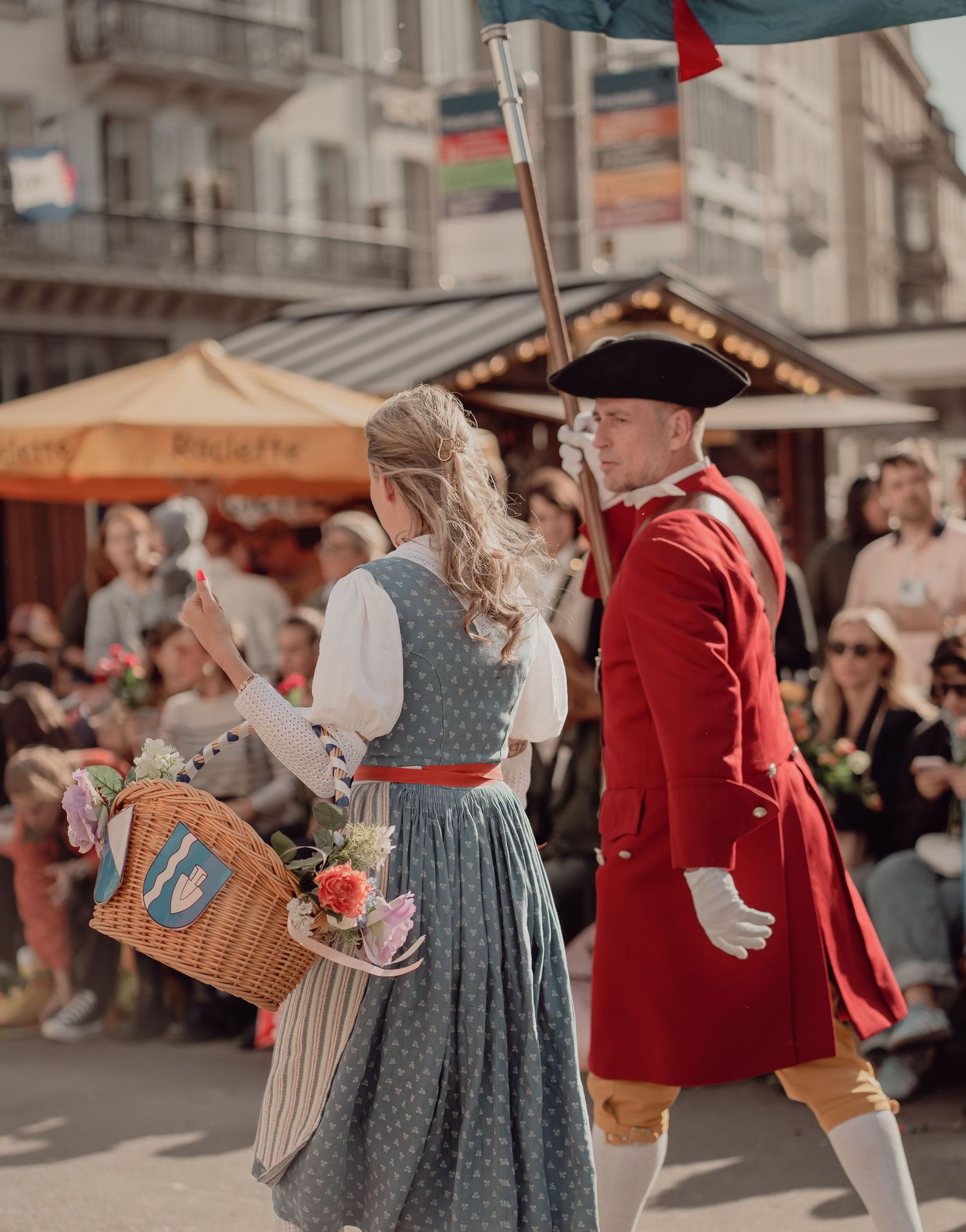 Woman in traditional dirndl dress with flower basket walks beside man in red historical uniform at a festive parade.