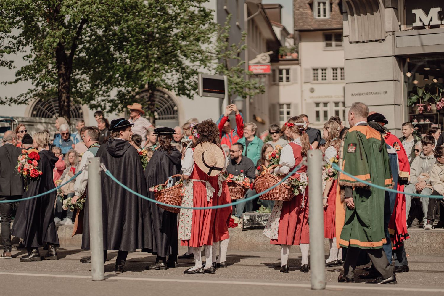 People in historical costumes gather at a festive outdoor parade with spectators watching nearby.