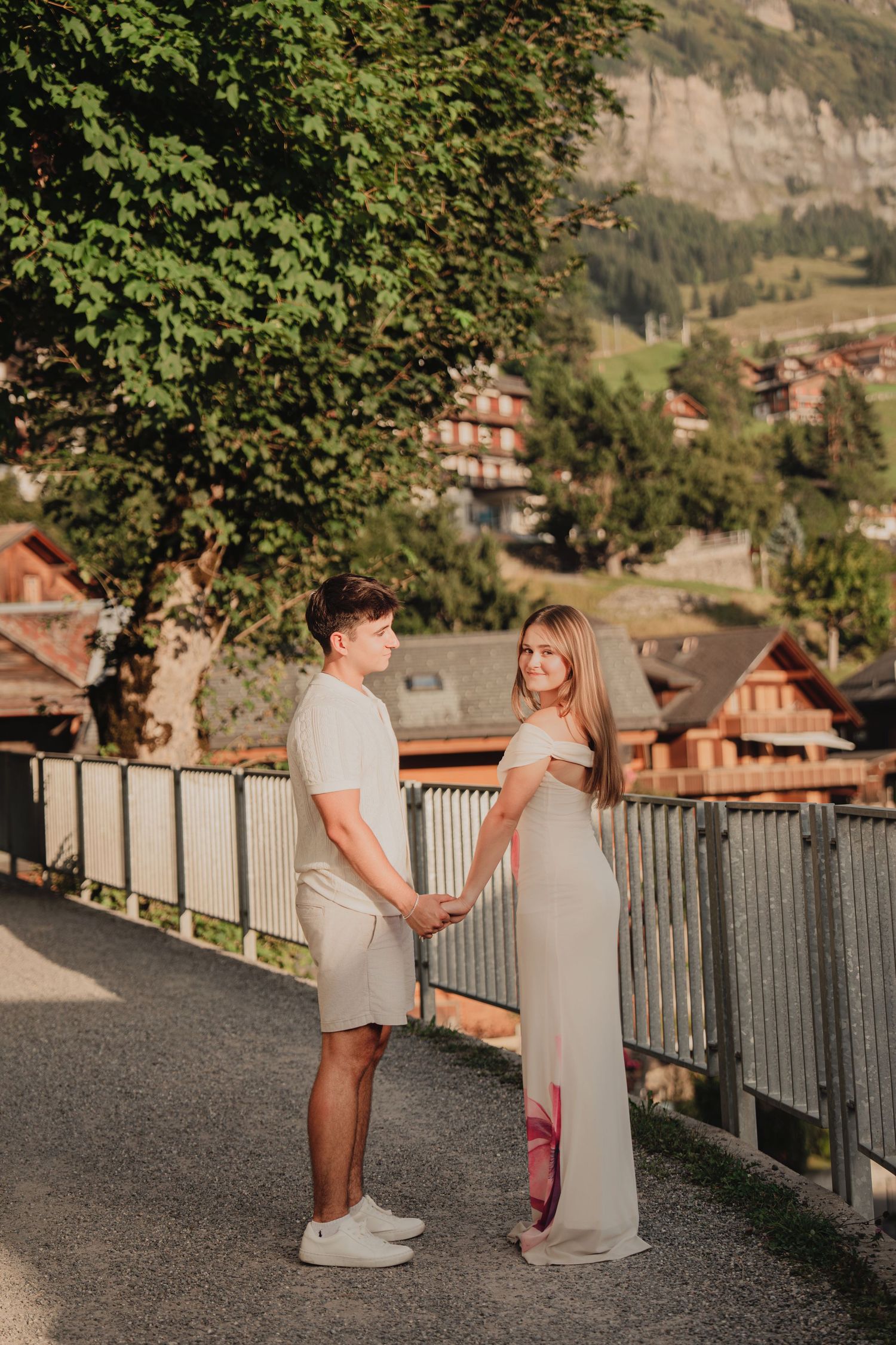 A couple holds hands facing each other on a bridge with a charming alpine town backdrop.
