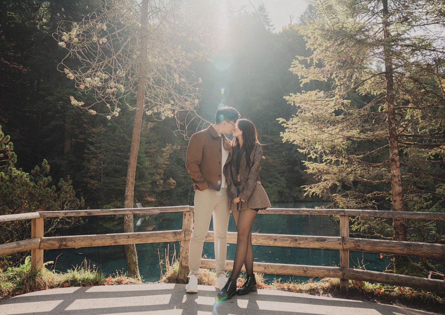Couple sharing a kiss on a wooden bridge bathed in golden sunlight with a blue lake in the background.