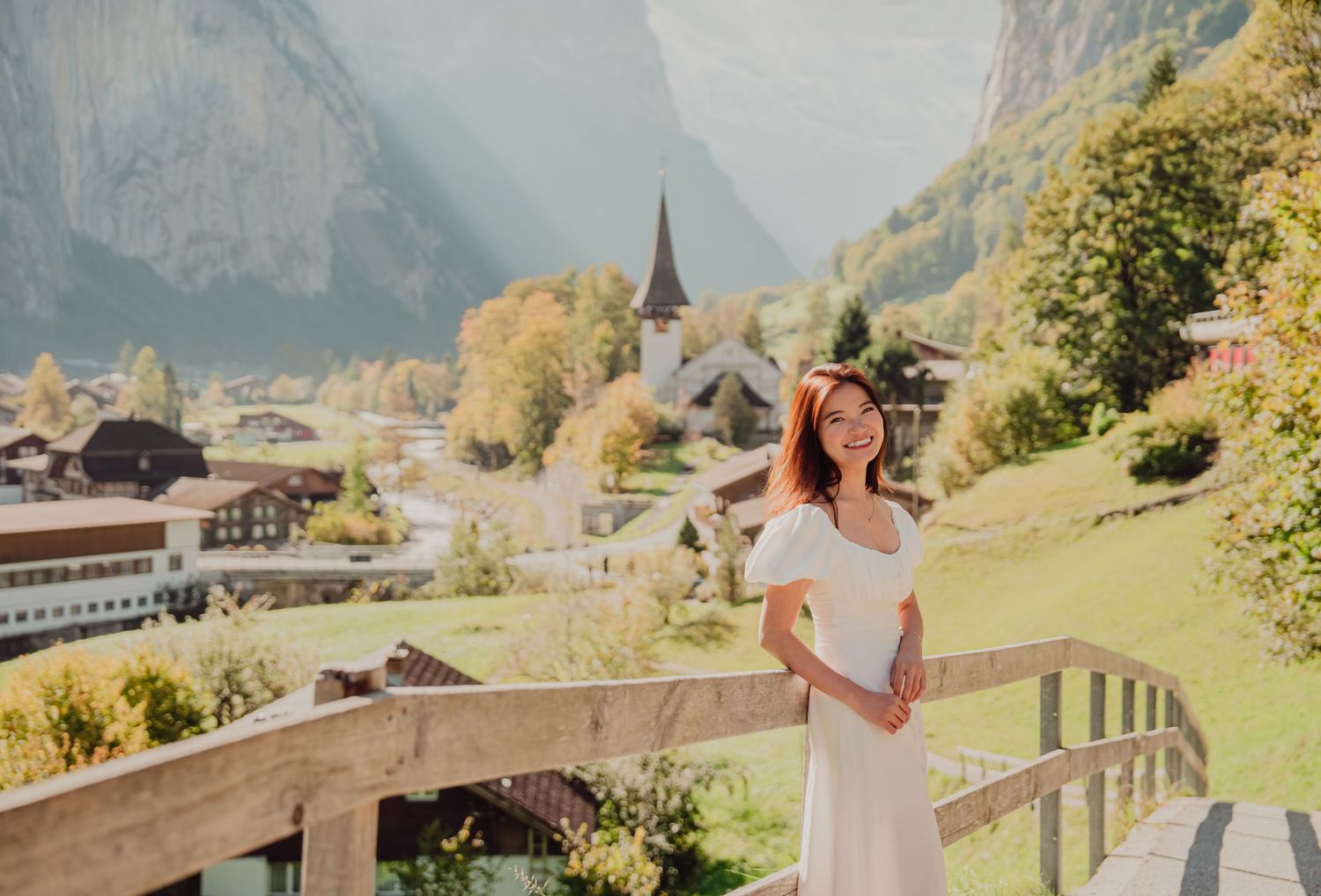 A cheerful woman in a white off-shoulder dress poses on a wooden bridge with a Swiss village backdrop.