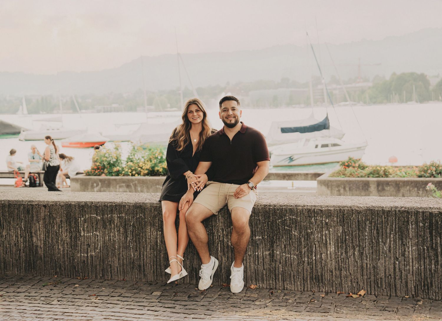 Couple sitting on a stone wall by a lake with sailboats, smiling at camera in Zurich, Switzerland.