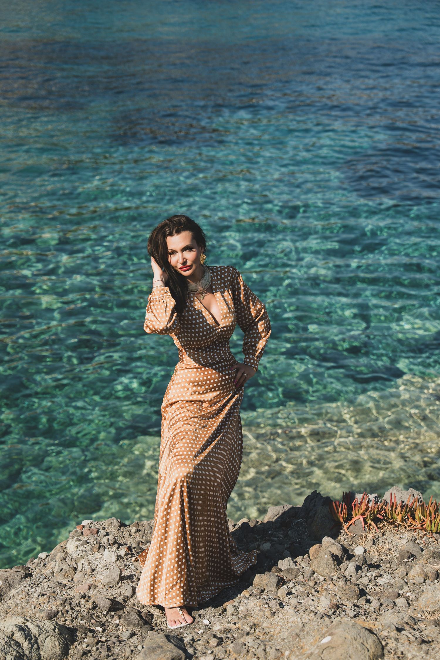 Stylish woman in long polka dot dress standing on rocks beside sparkling blue sea.