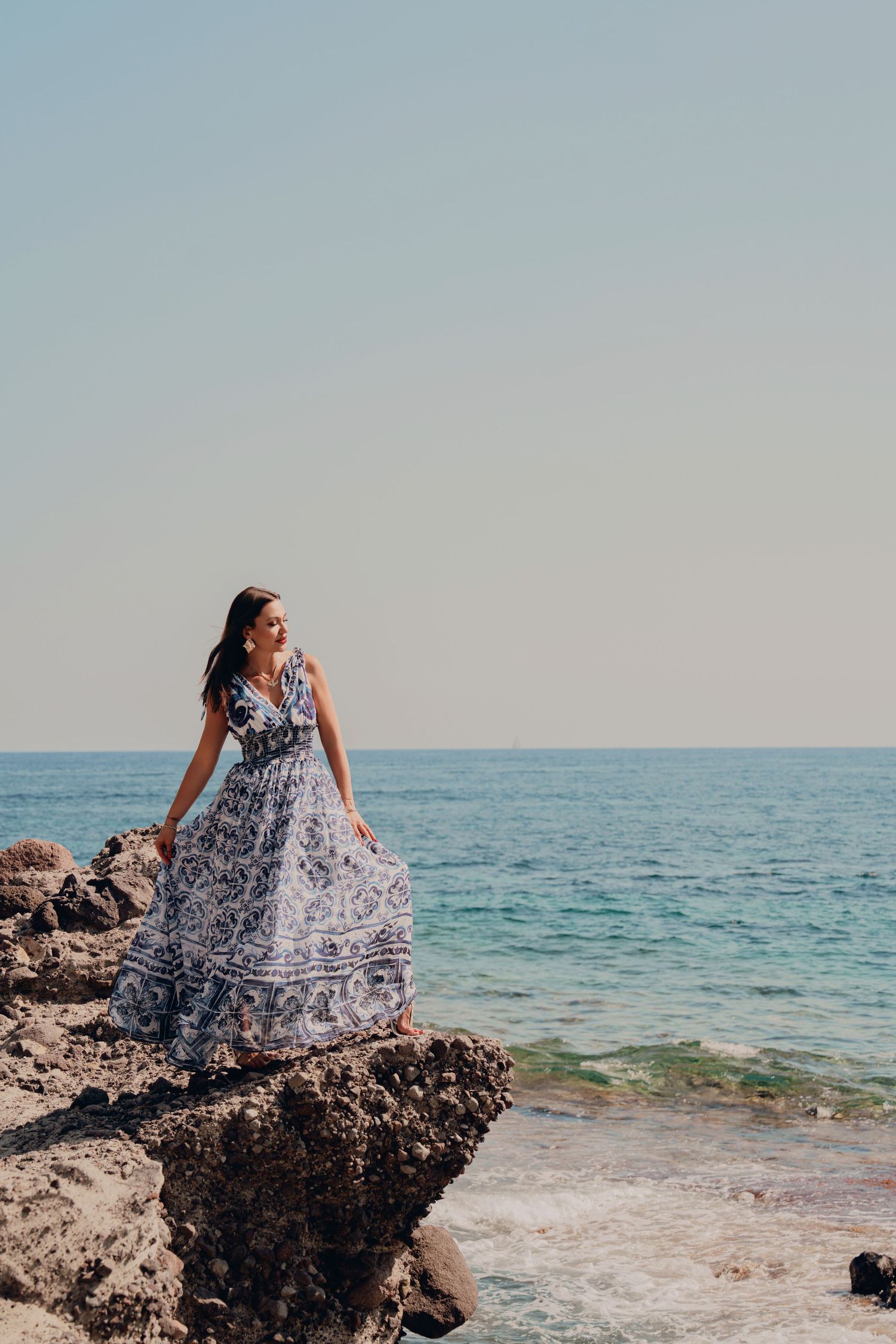 Woman in blue and white patterned maxi dress posing on rocky seaside with ocean view.
