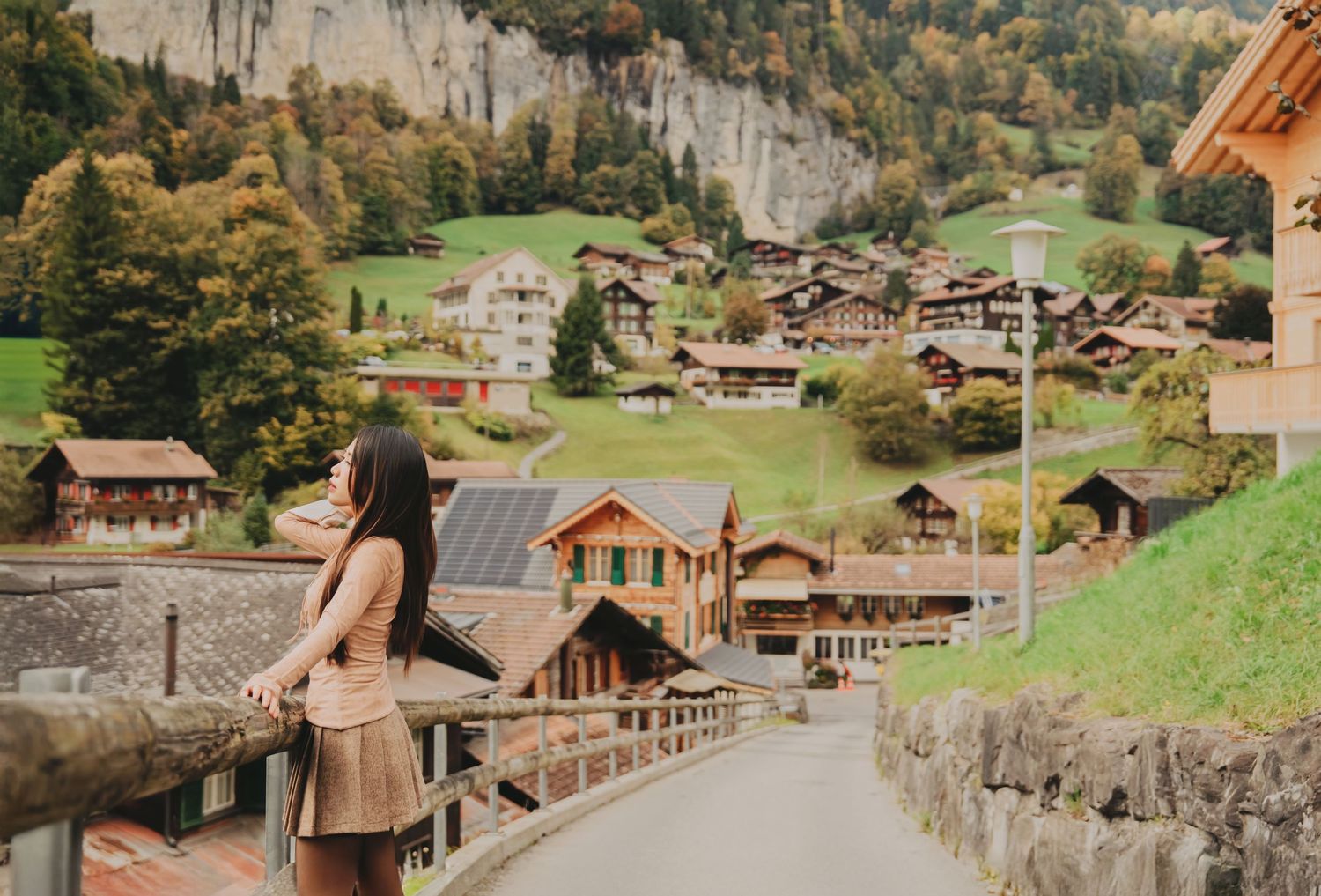 Woman in skirt leaning on bridge railing overlooking a charming Swiss alpine village with colorful autumn hillsides.