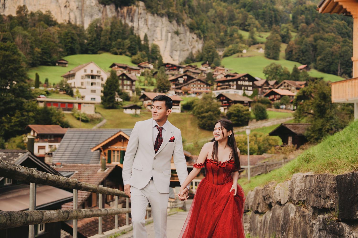 Couple walking hand in hand on a path with charming Swiss chalet village and cliffs behind them.