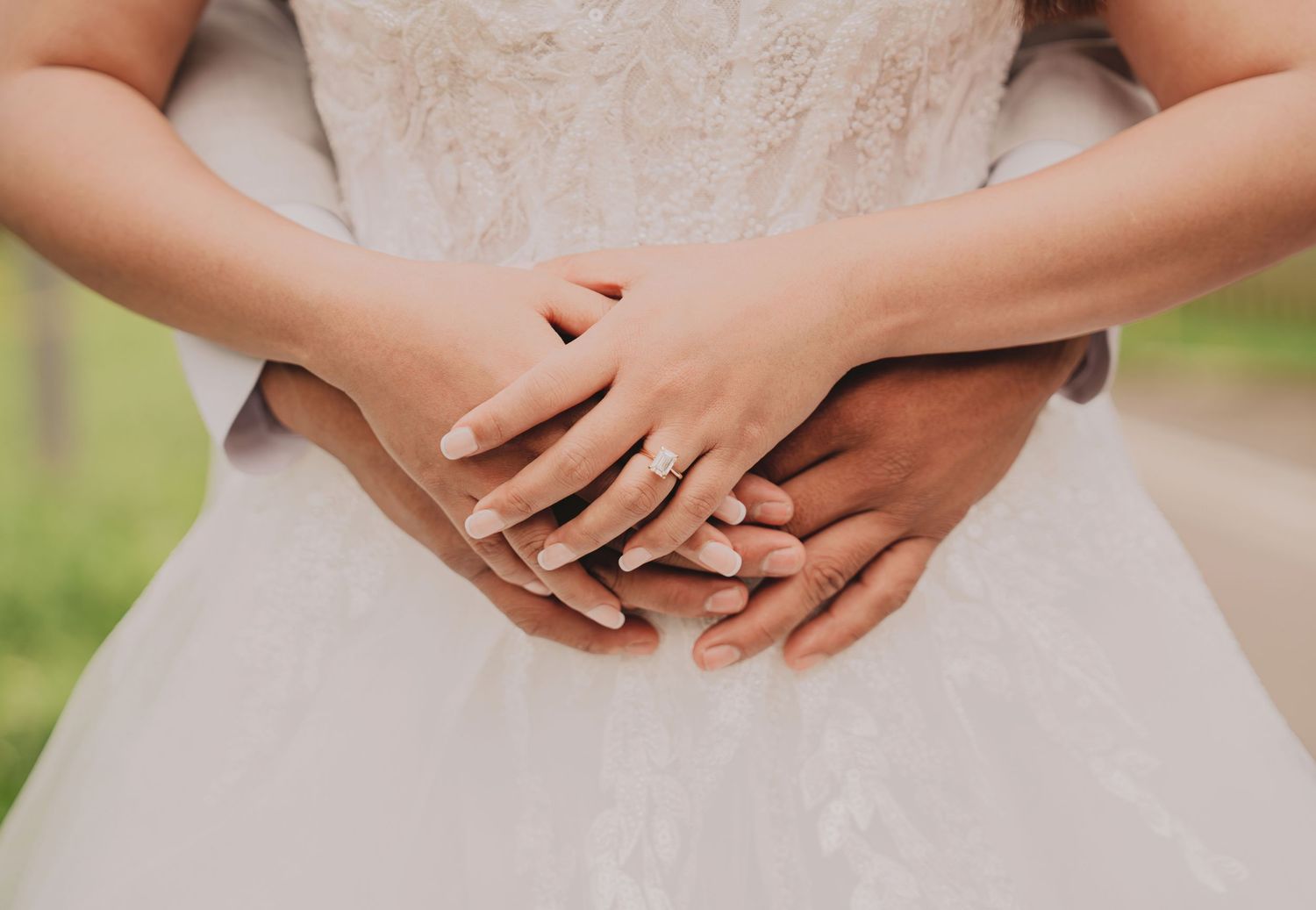 Bride and groom holding hands together displaying wedding rings against white lace bridal gown.