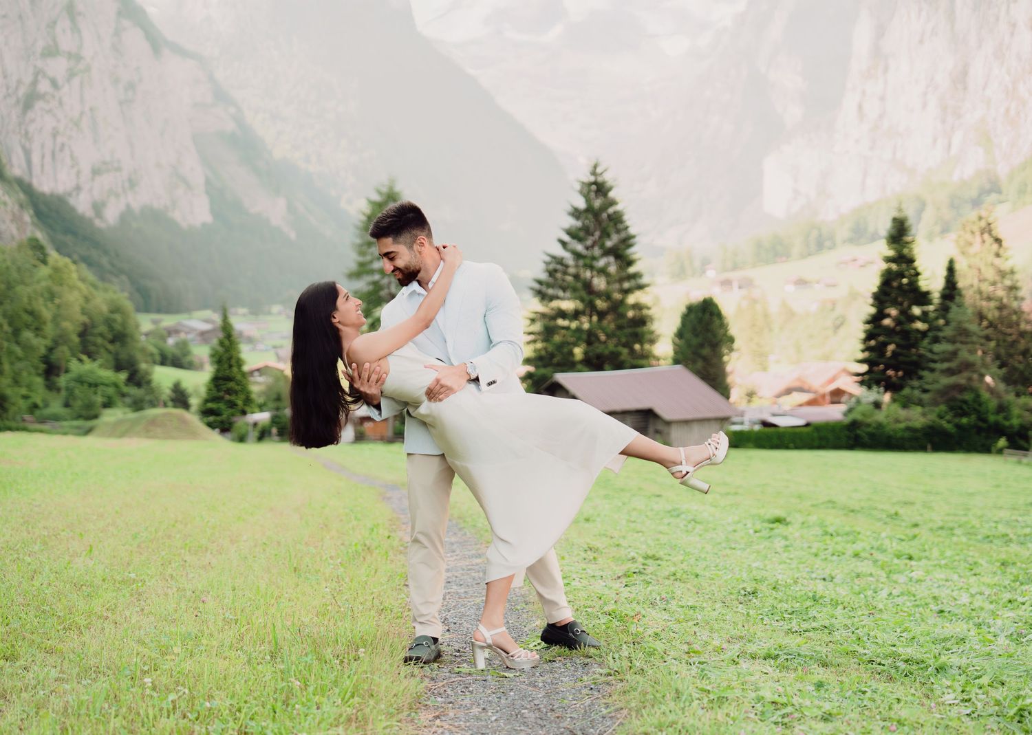 Couple sharing a romantic dip embrace on green meadow with dramatic Swiss mountain cliffs.