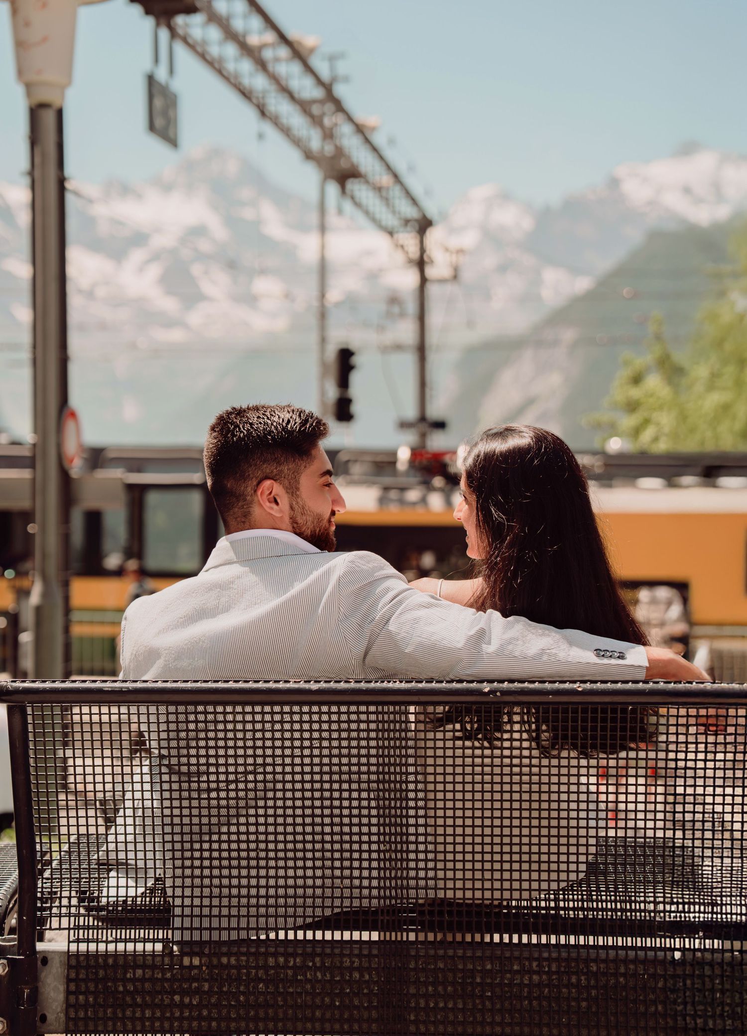 Romantic couple sitting on a bench from behind with Swiss Alps and yellow train in background.