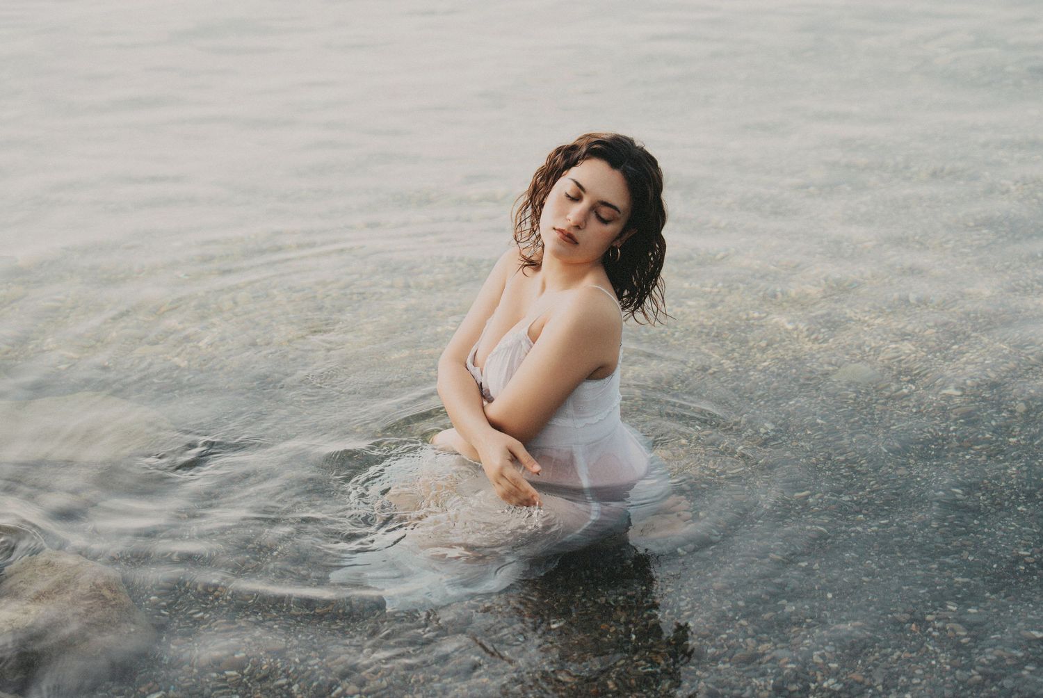 Woman in wet white dress sitting in shallow water by the shore.