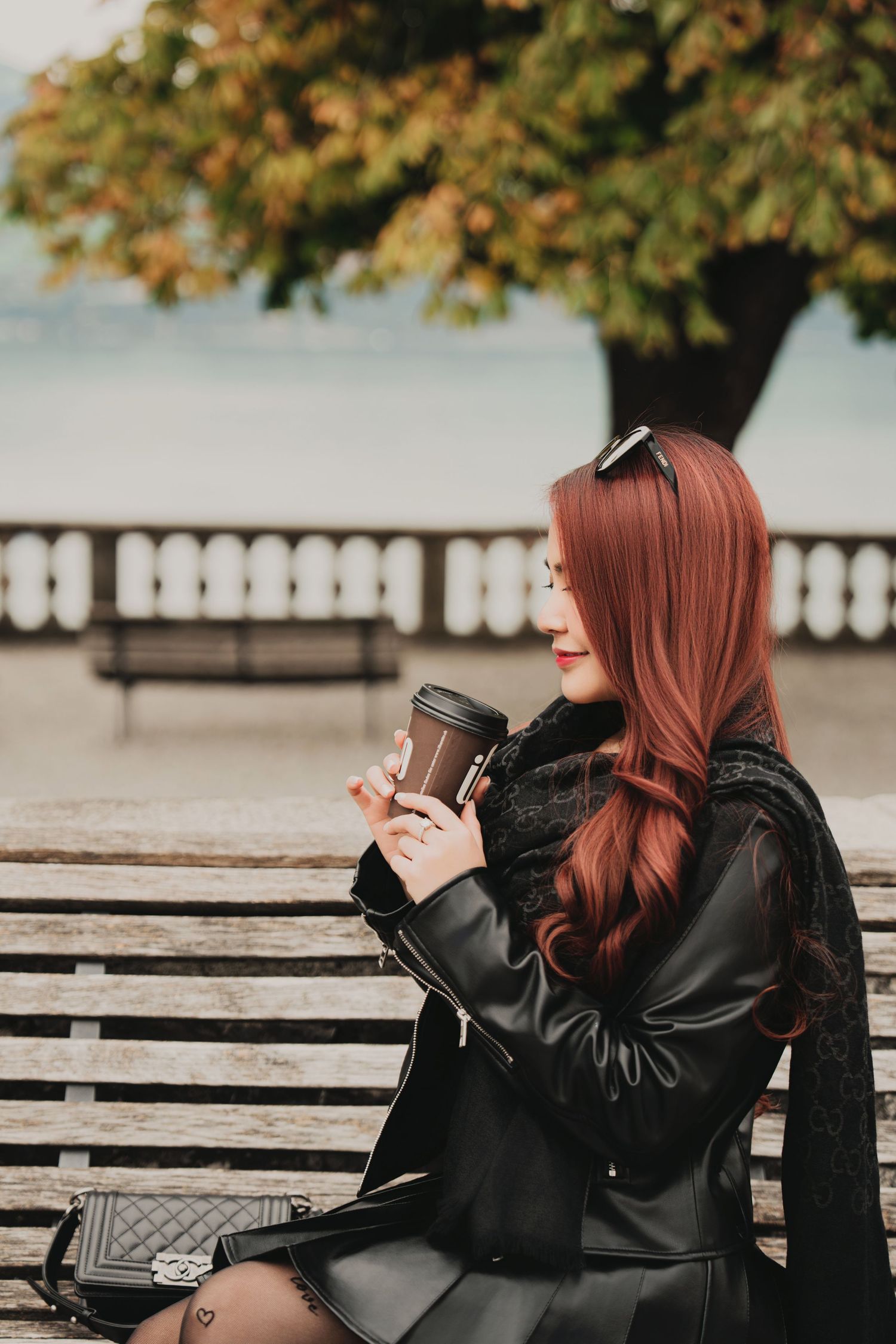 Stylish red-haired woman in black leather outfit sits on wooden bench holding coffee cup near autumn lakeside park.
