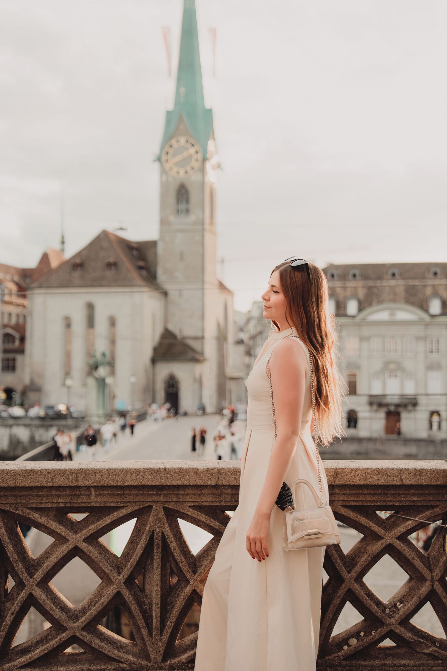 Young woman in elegant cream dress poses on historic Munsterbrucke bridge with Fraumunster Church, Zurich.