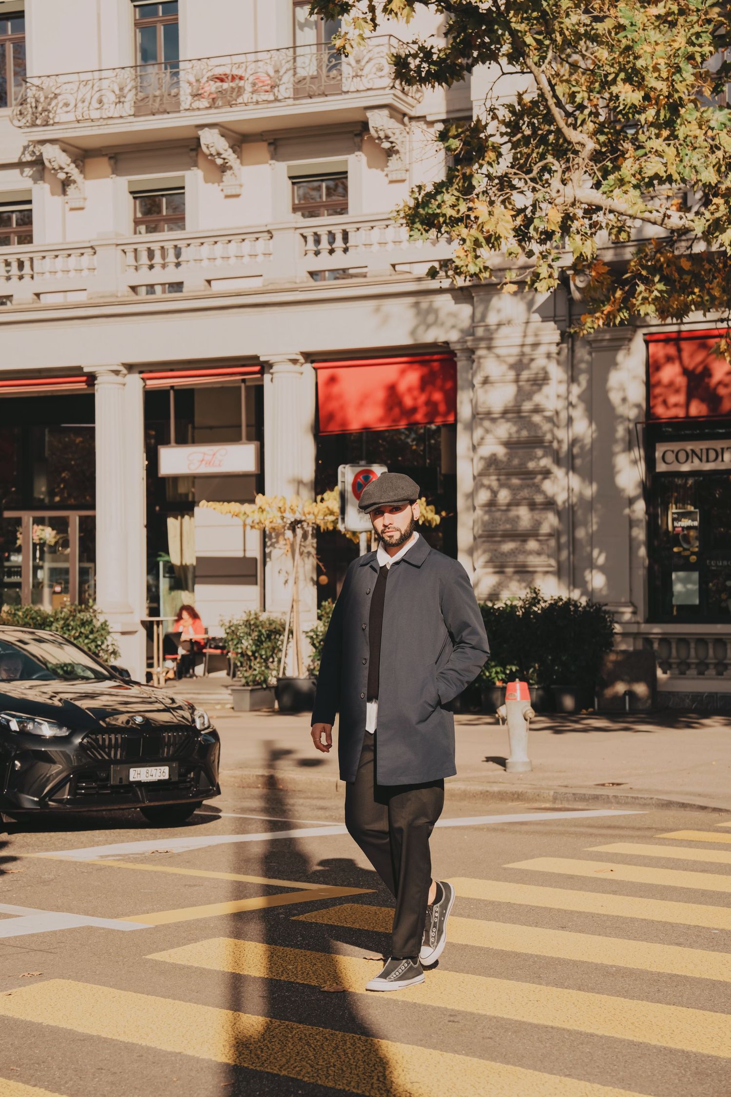 Bearded man in dark coat and flat cap crosses a sunny urban street with elegant buildings behind.