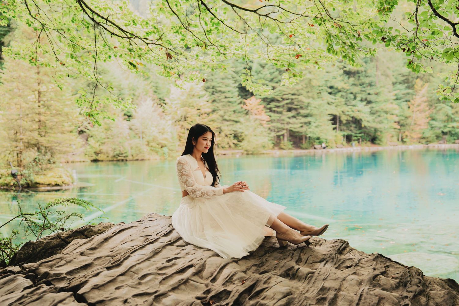Woman in white dress sits peacefully on rock by serene turquoise lake in forest.