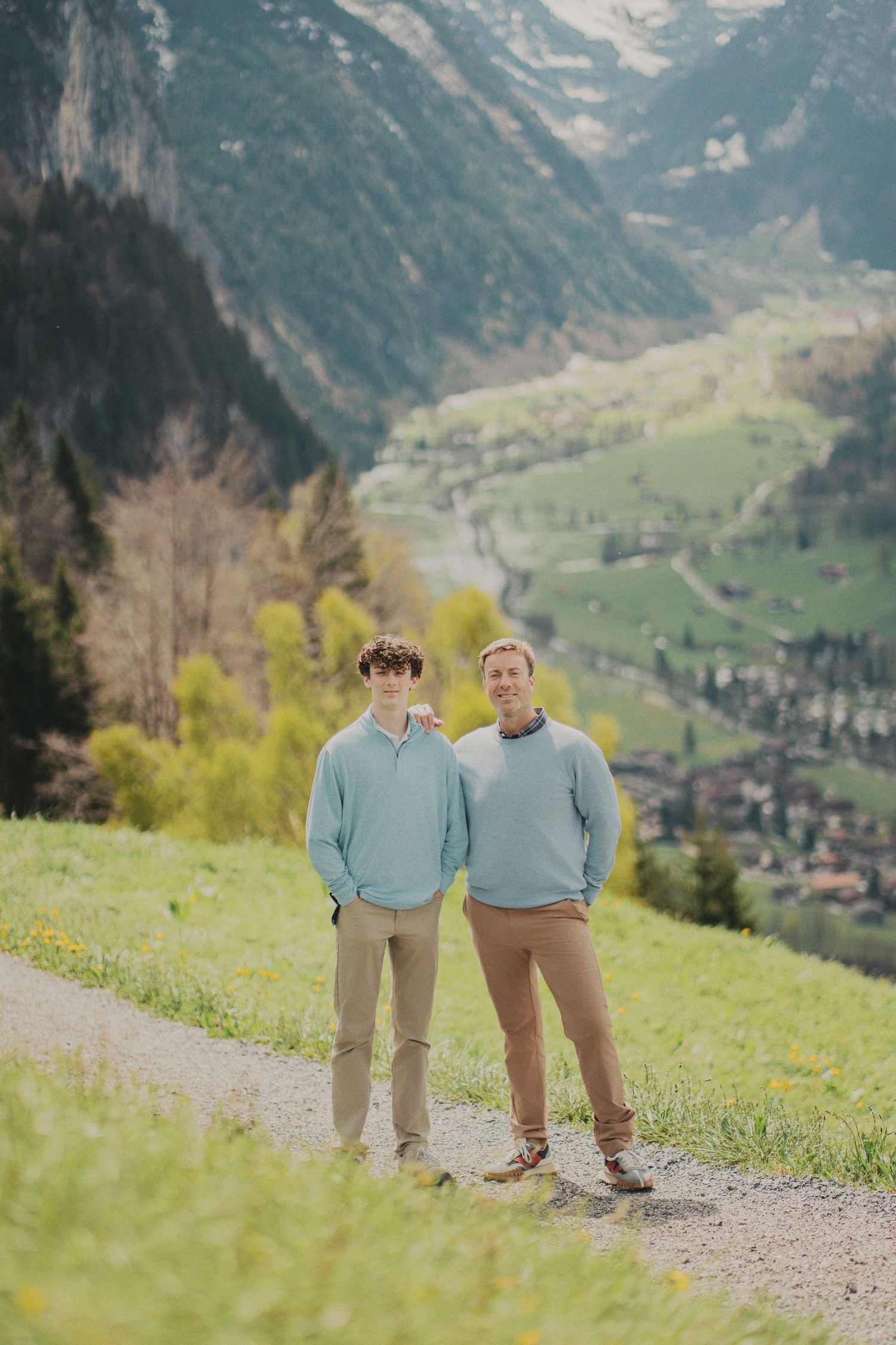 Father and teenage son in matching blue pullovers standing on a hillside path with green Swiss valley below.