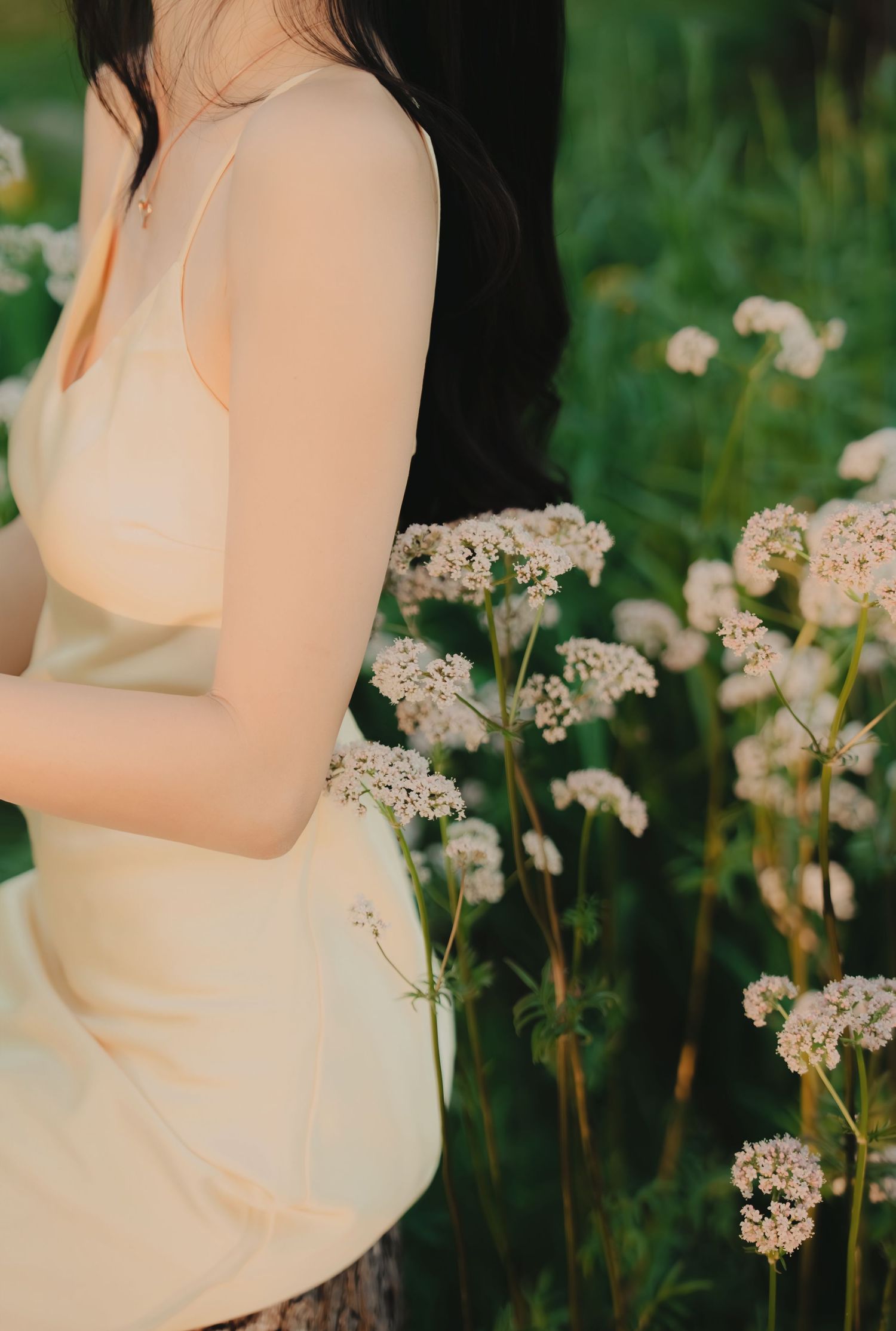 Young woman in cream slip dress beside white wildflowers in a lush green meadow at sunset.