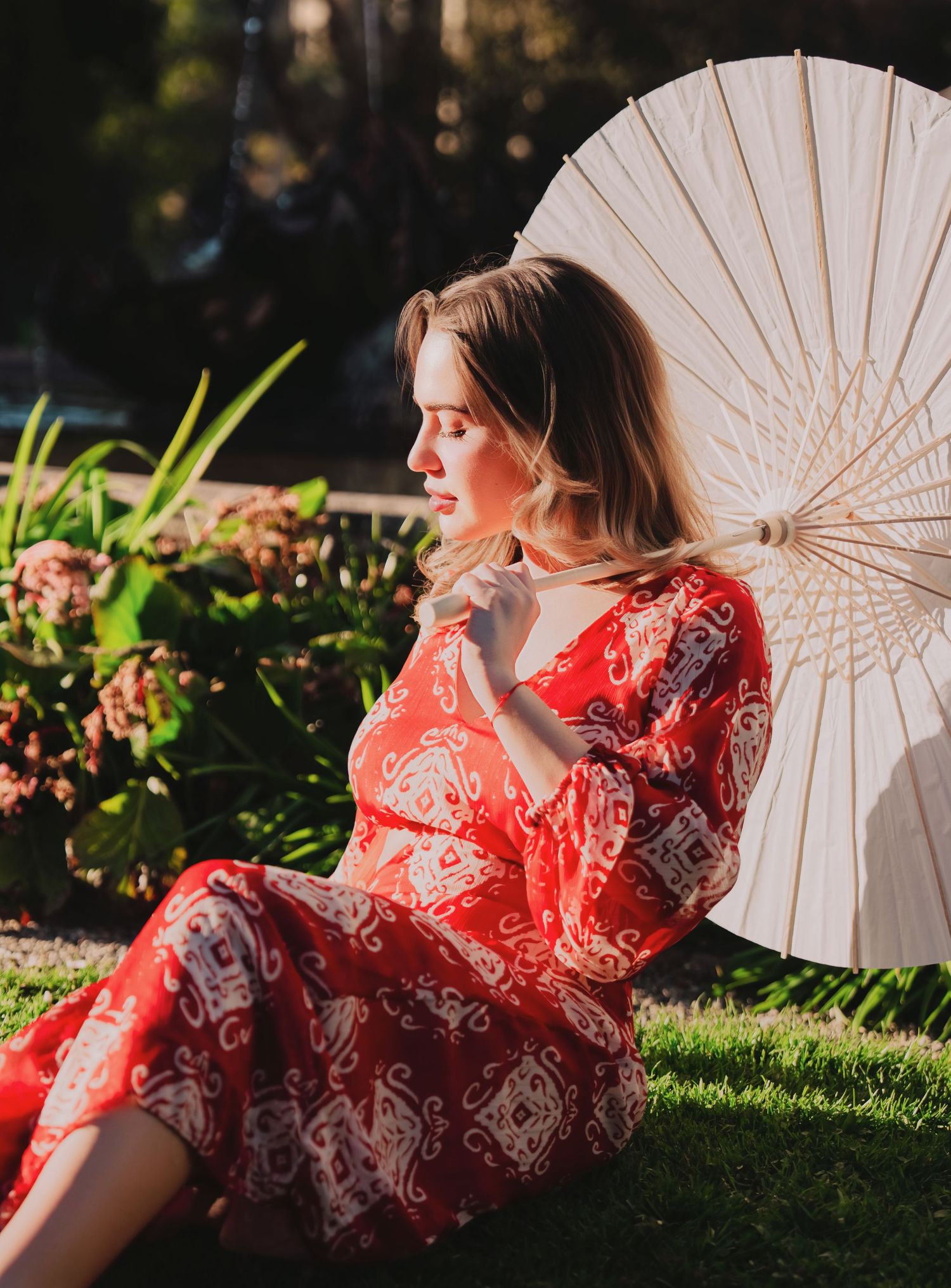 Young woman in red paisley dress relaxing on grass with white parasol in sunlight.