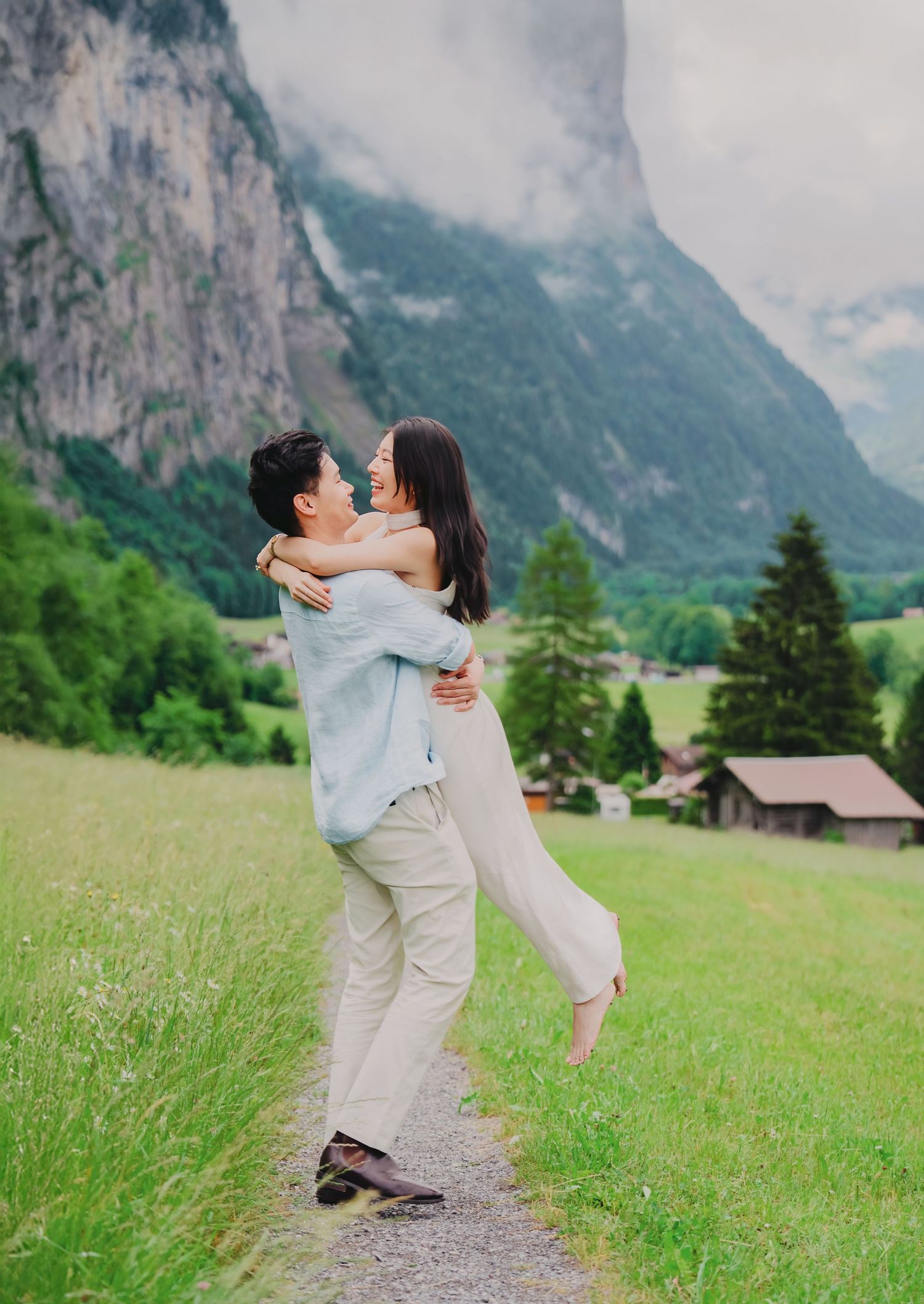 Man lifting woman in his arms on a green alpine meadow with dramatic mountain cliffs behind.