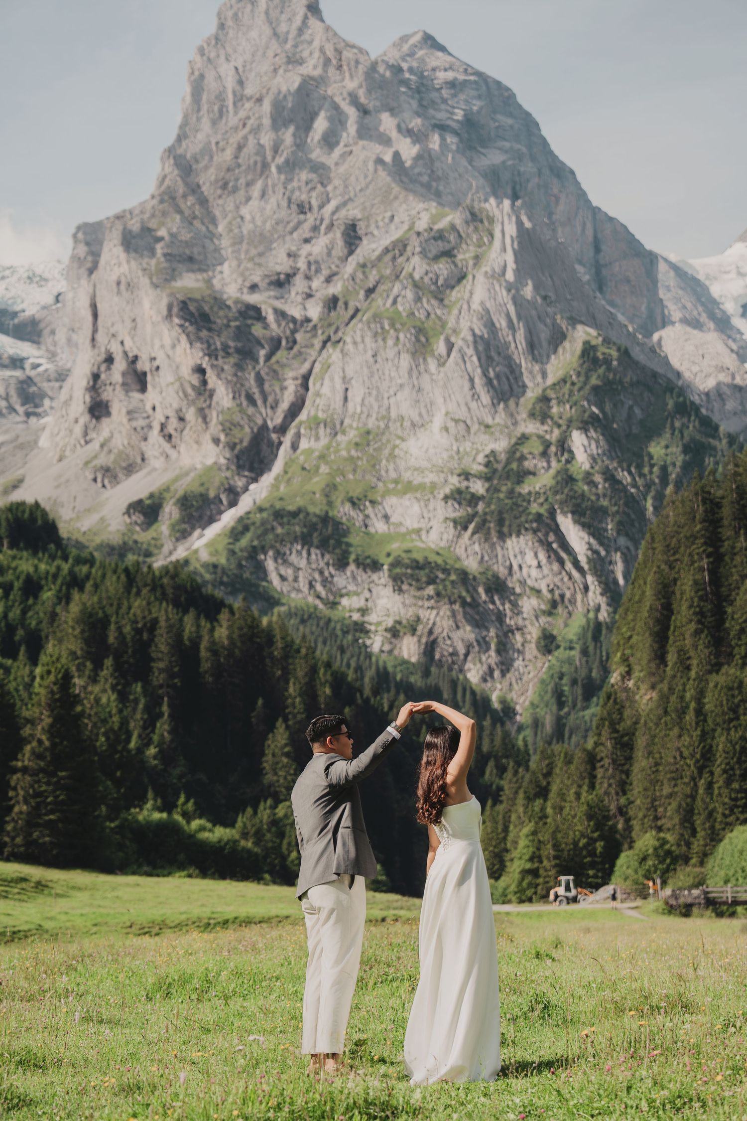 Groom twirls bride in white dress on green meadow with towering Swiss Alps in background.