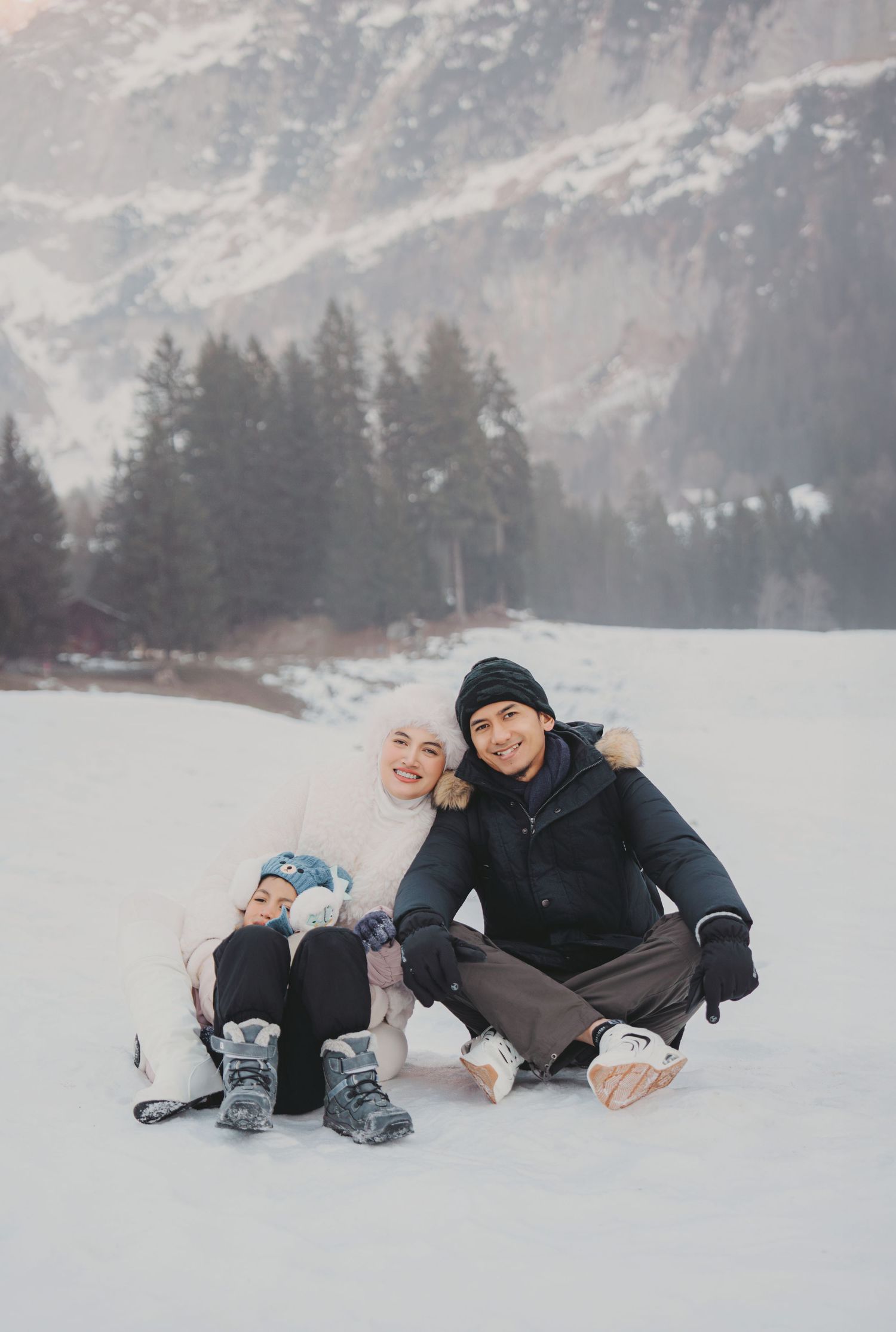 Happy family of three sitting together on snow with mountain forest backdrop in winter.