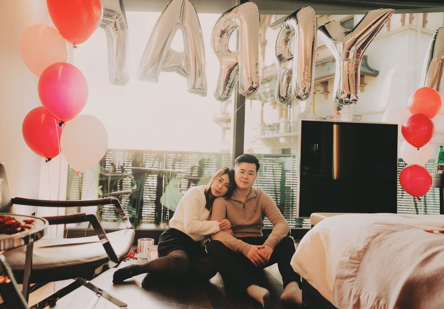 Asian couple sitting together in a decorated room with MARRY ME balloons and red heart balloons.