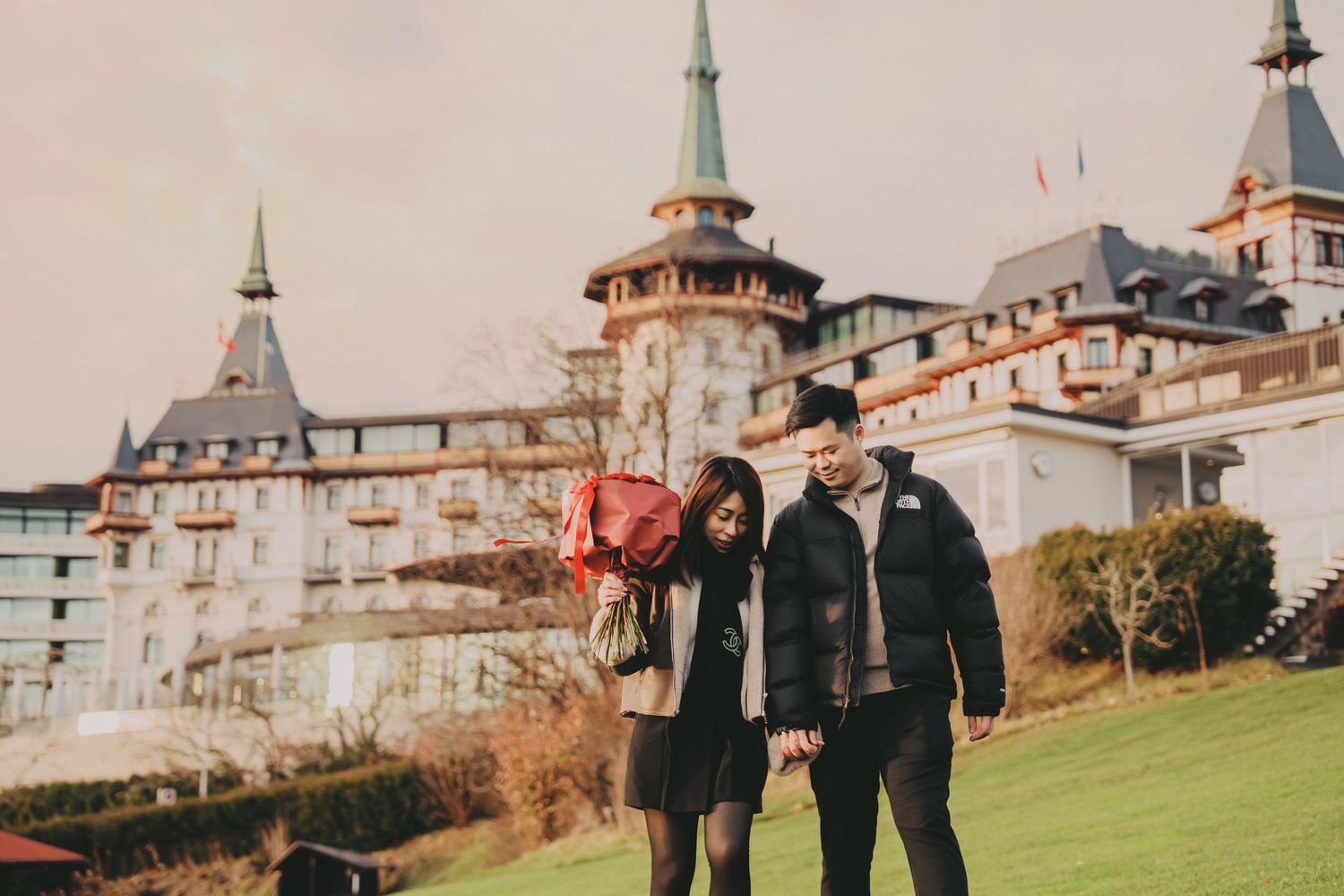 Couple walking hand in hand on green lawn with bouquet of red roses before grand European castle.