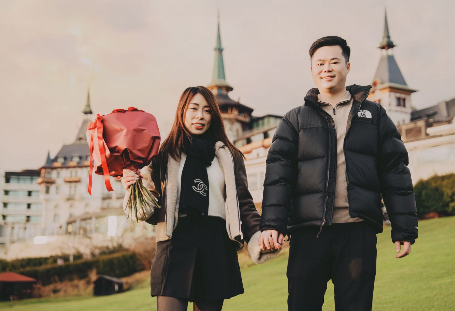 Asian couple walking hand in hand with red bouquet near historic castle hotel.