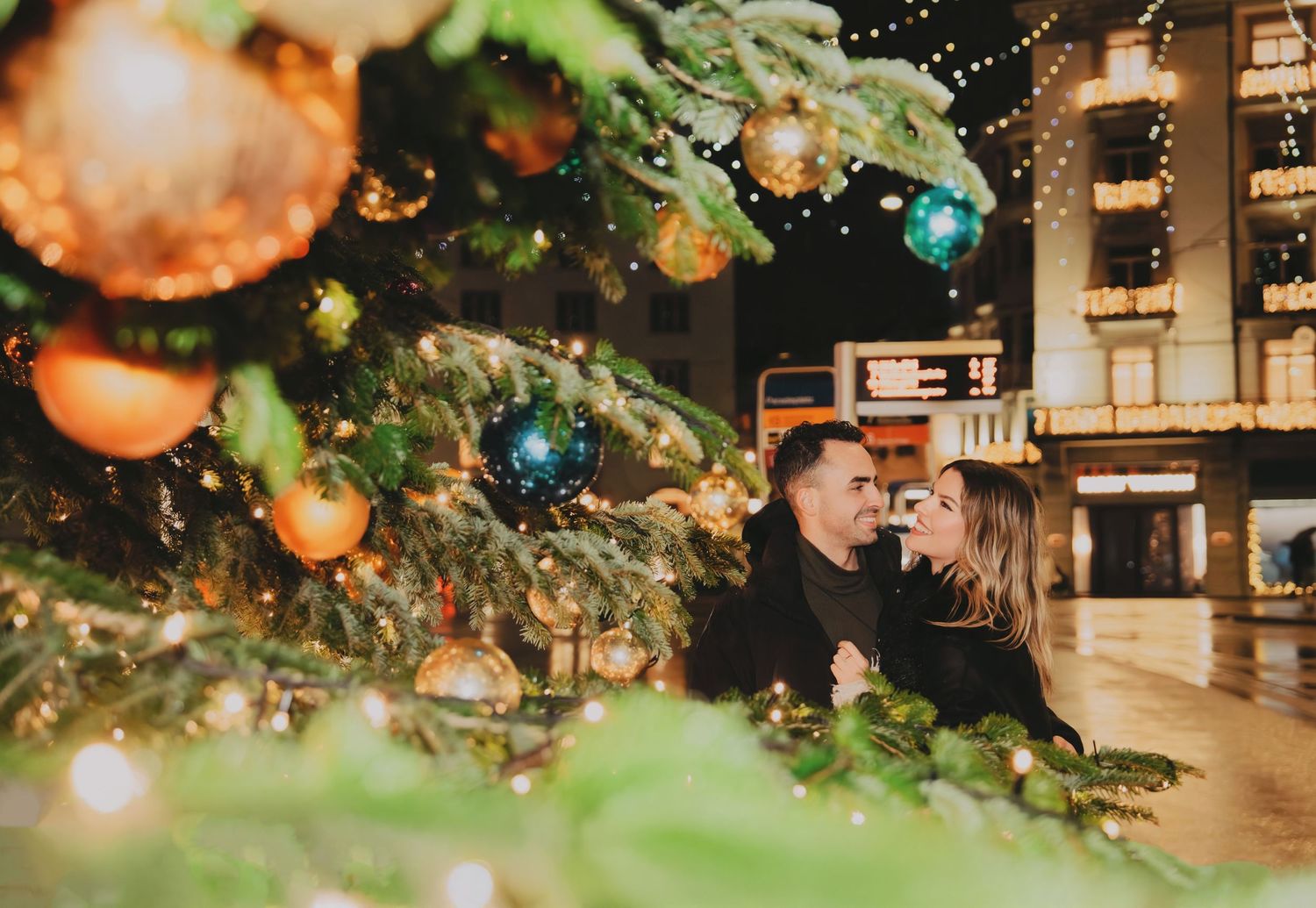 Couple sharing a romantic moment framed by Christmas tree ornaments and bokeh lights.