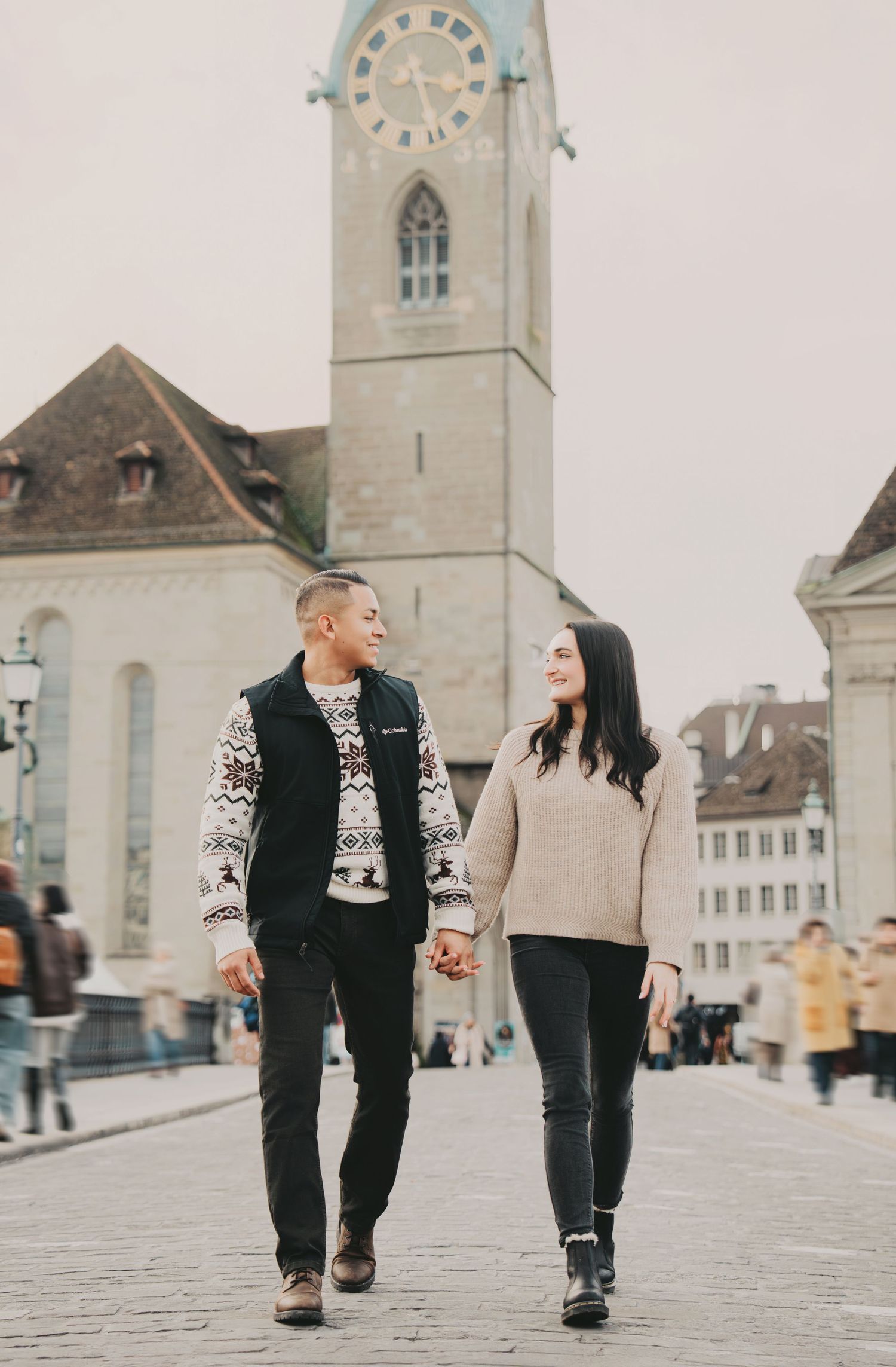 Couple walking and smiling at each other on a cobblestone square with Zurich's Fraumunster Church behind.