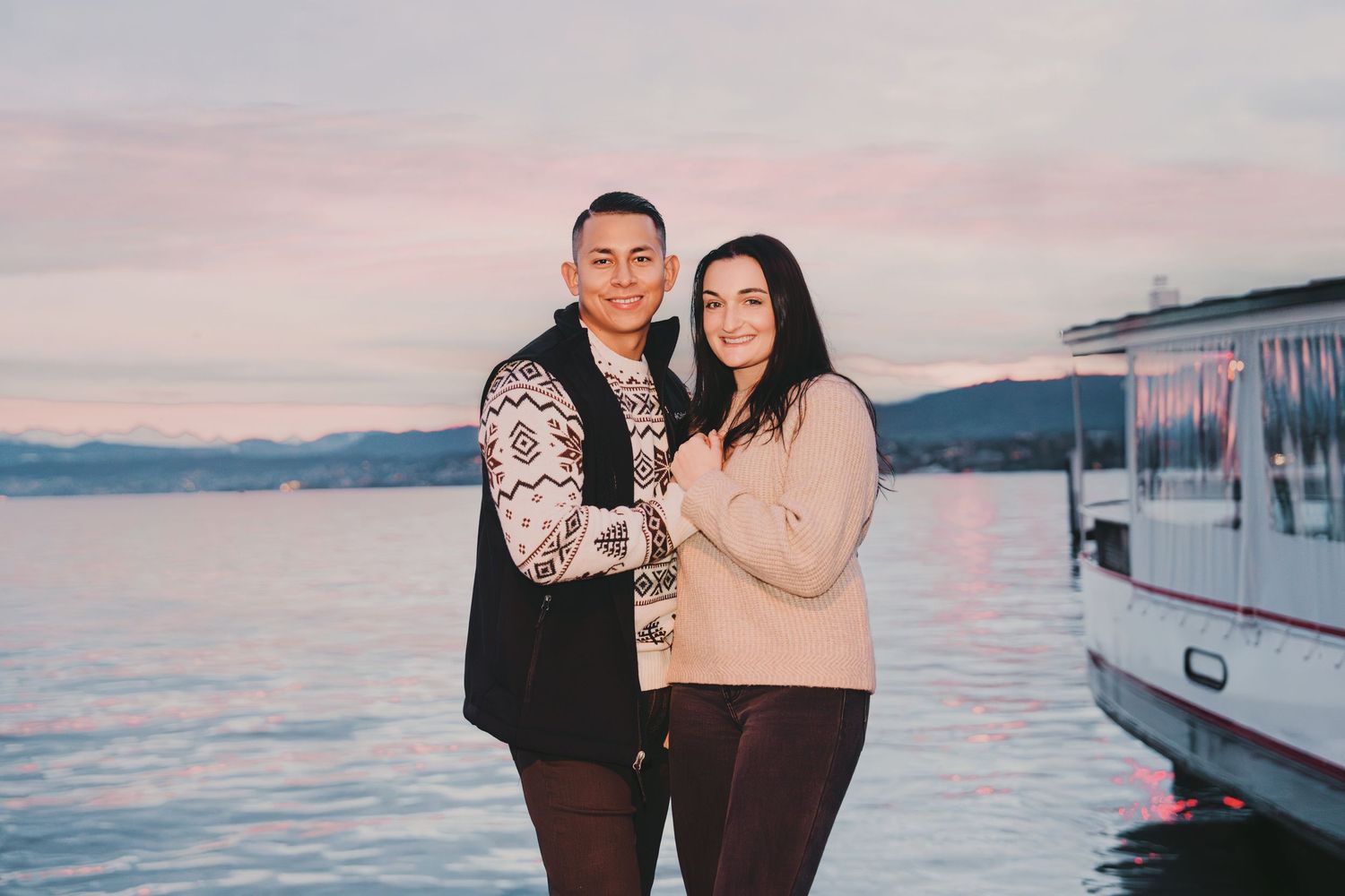 Romantic couple embracing on a lakeside dock at sunset with pastel pink sky.