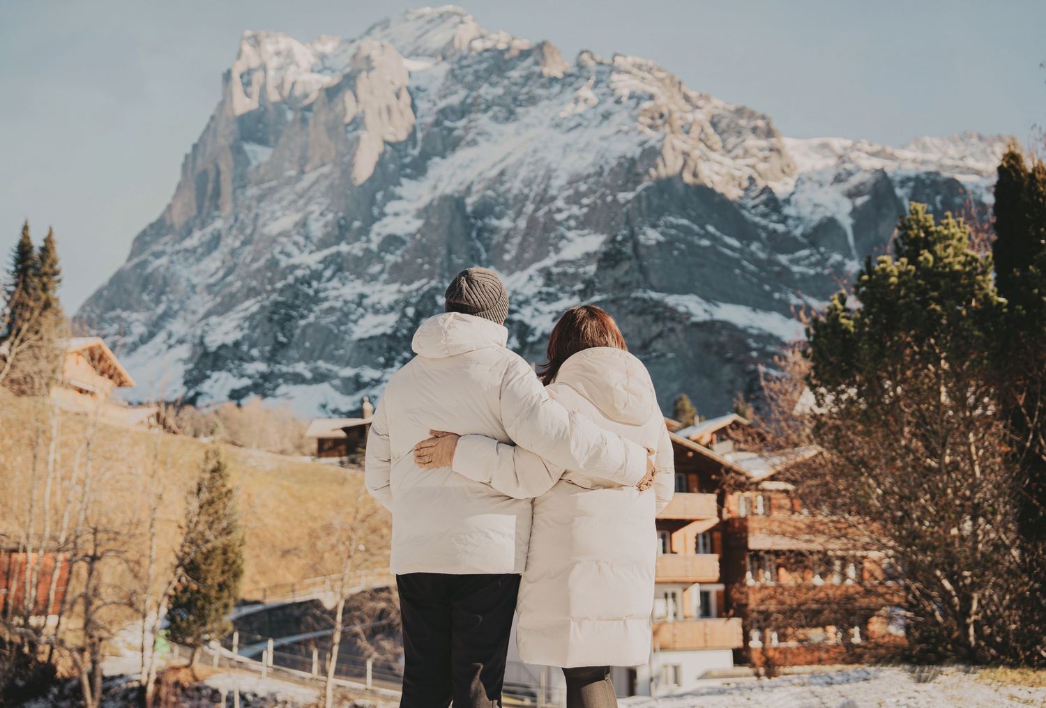 Couple with arms around each other admiring snow-capped mountain and sunny alpine village scenery.