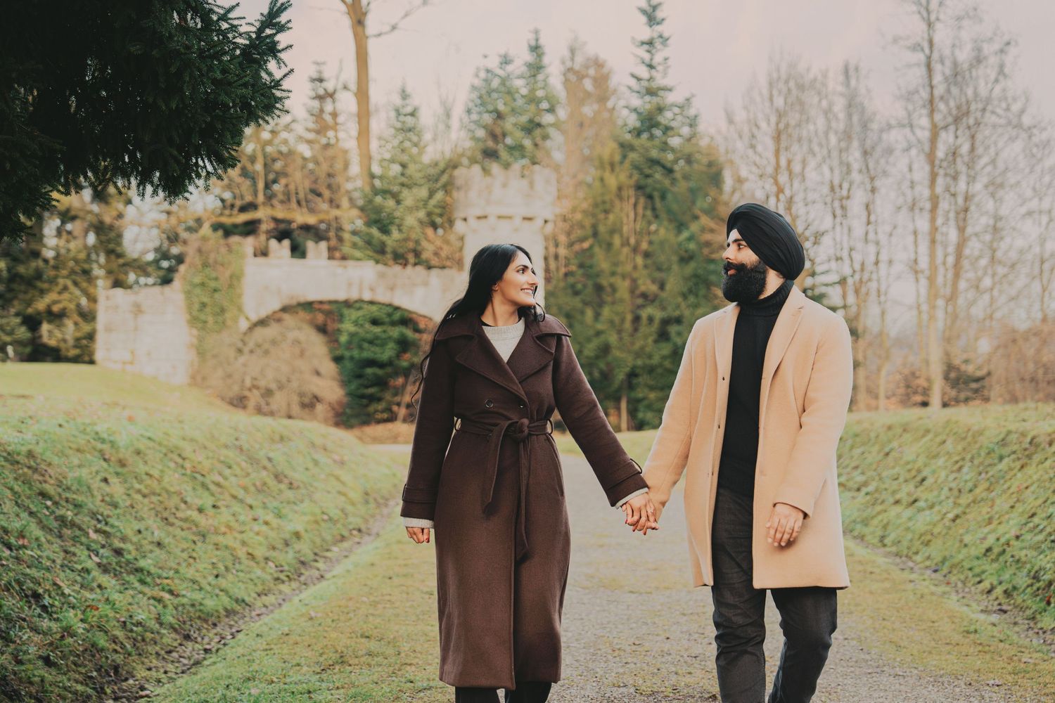 Couple holding hands and smiling at each other on a path with a stone bridge in the background.