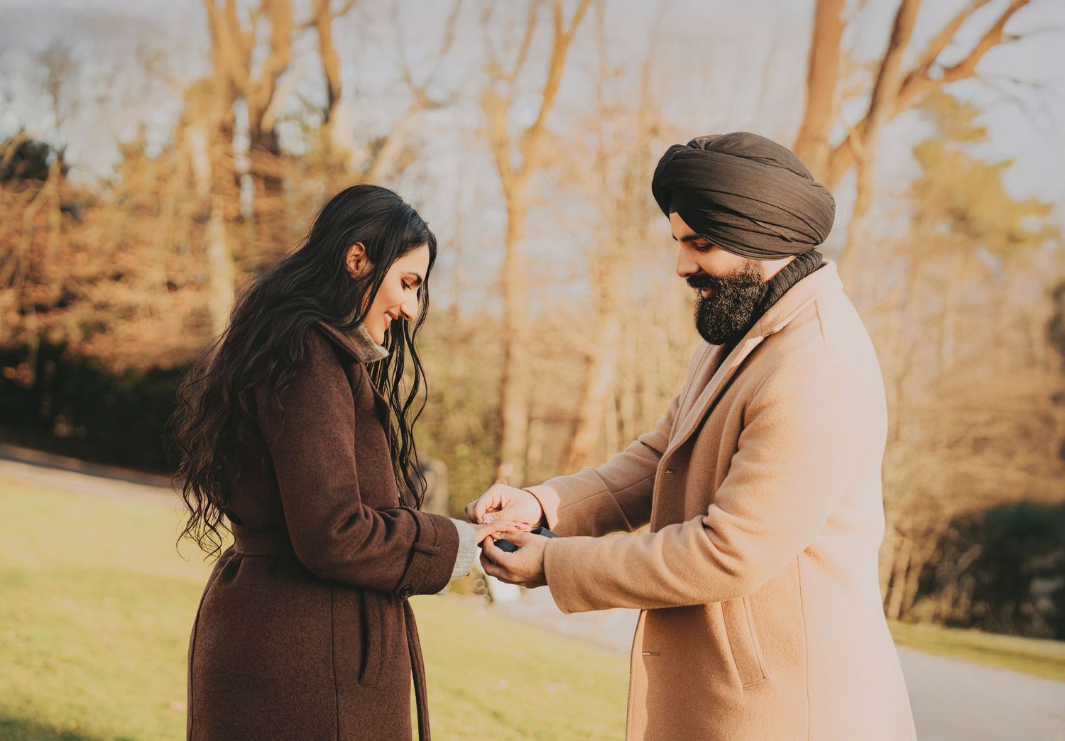 Sikh man in beige coat presenting engagement ring to woman in brown coat in autumn park.