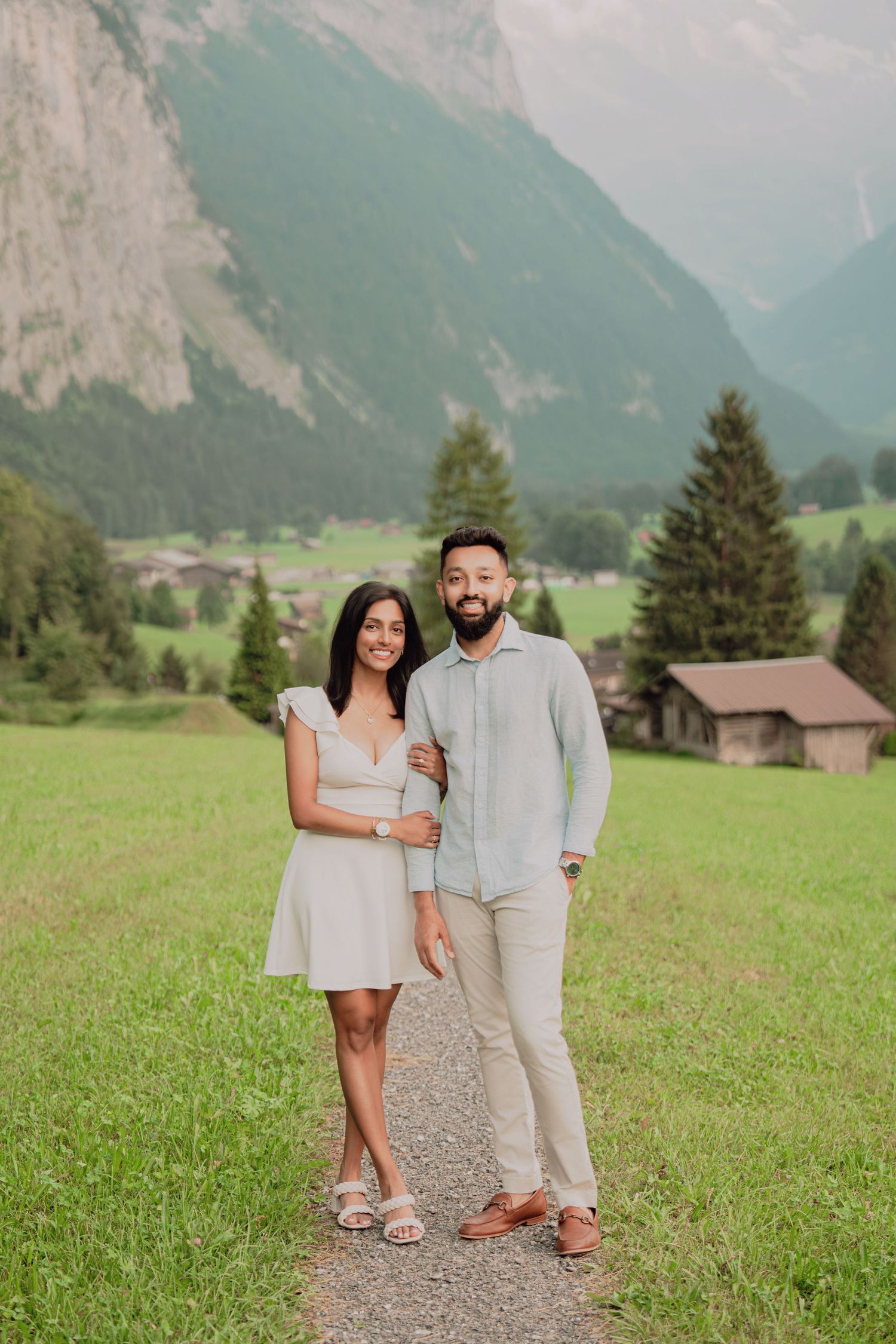 Engaged couple posing together on a scenic alpine path surrounded by green meadows and mountains.