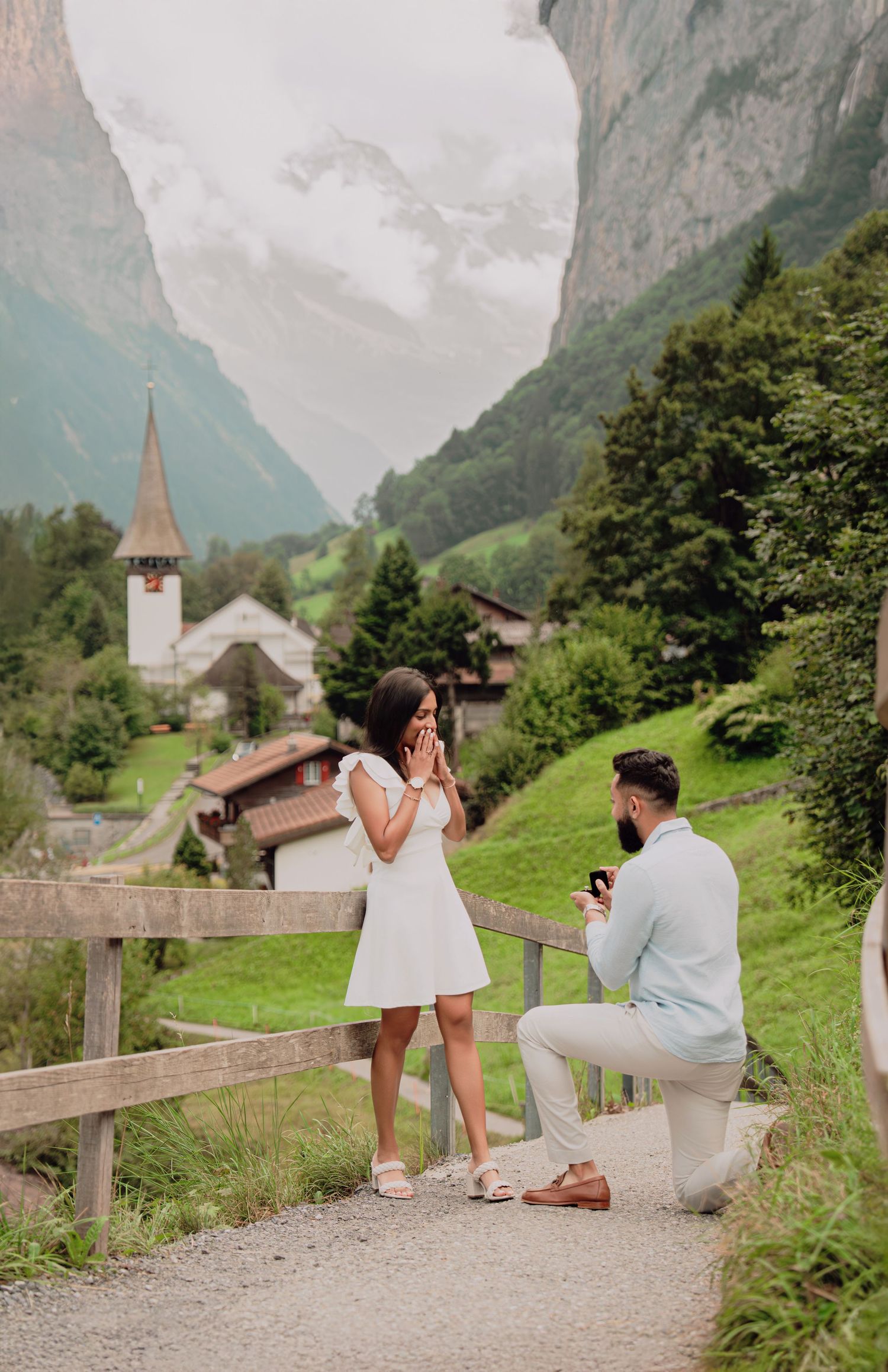 A man proposes on one knee to a tearful woman on a scenic Swiss Alpine path near a village church.