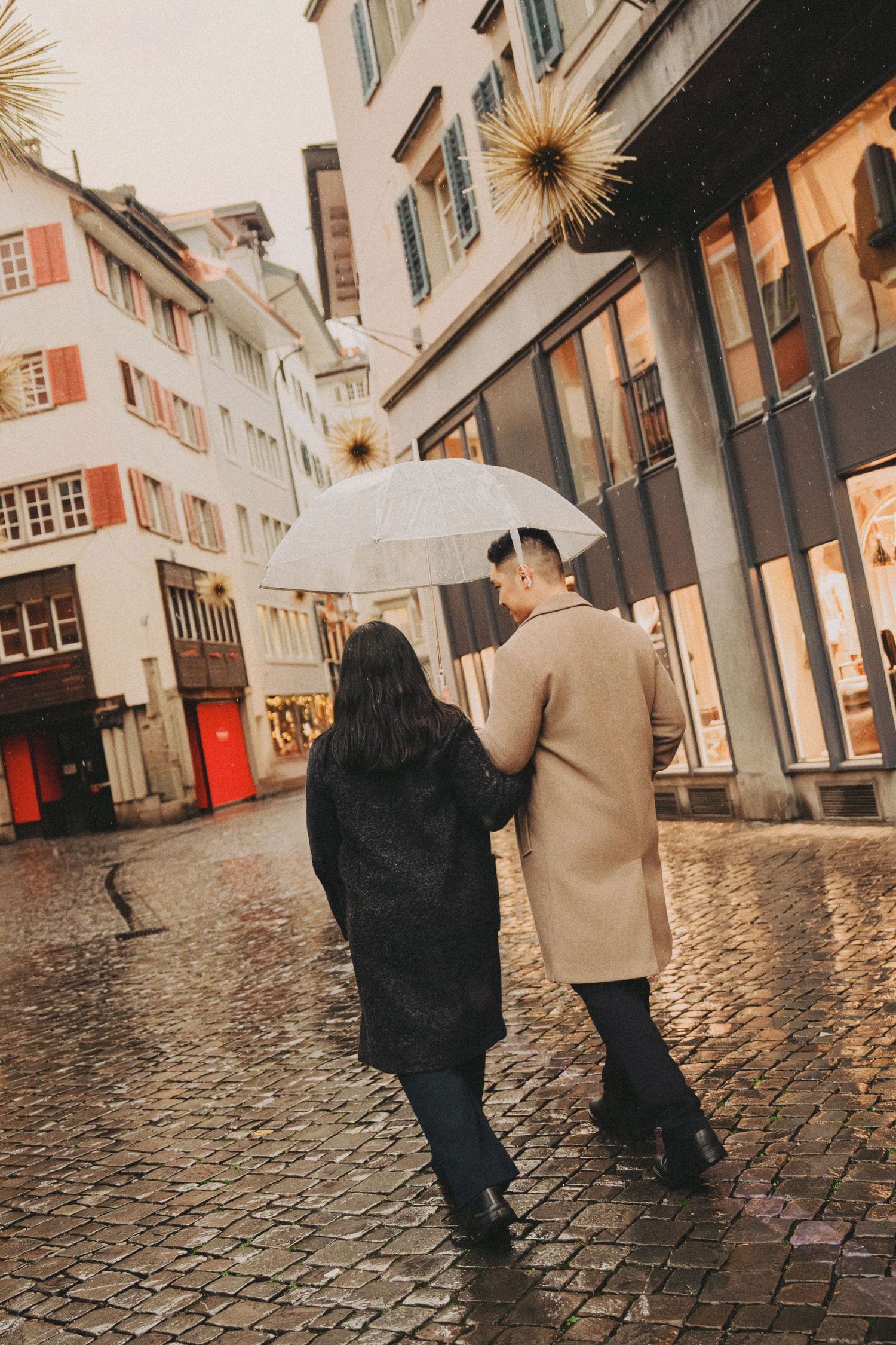 Couple walking away under umbrella on wet cobblestone European street.