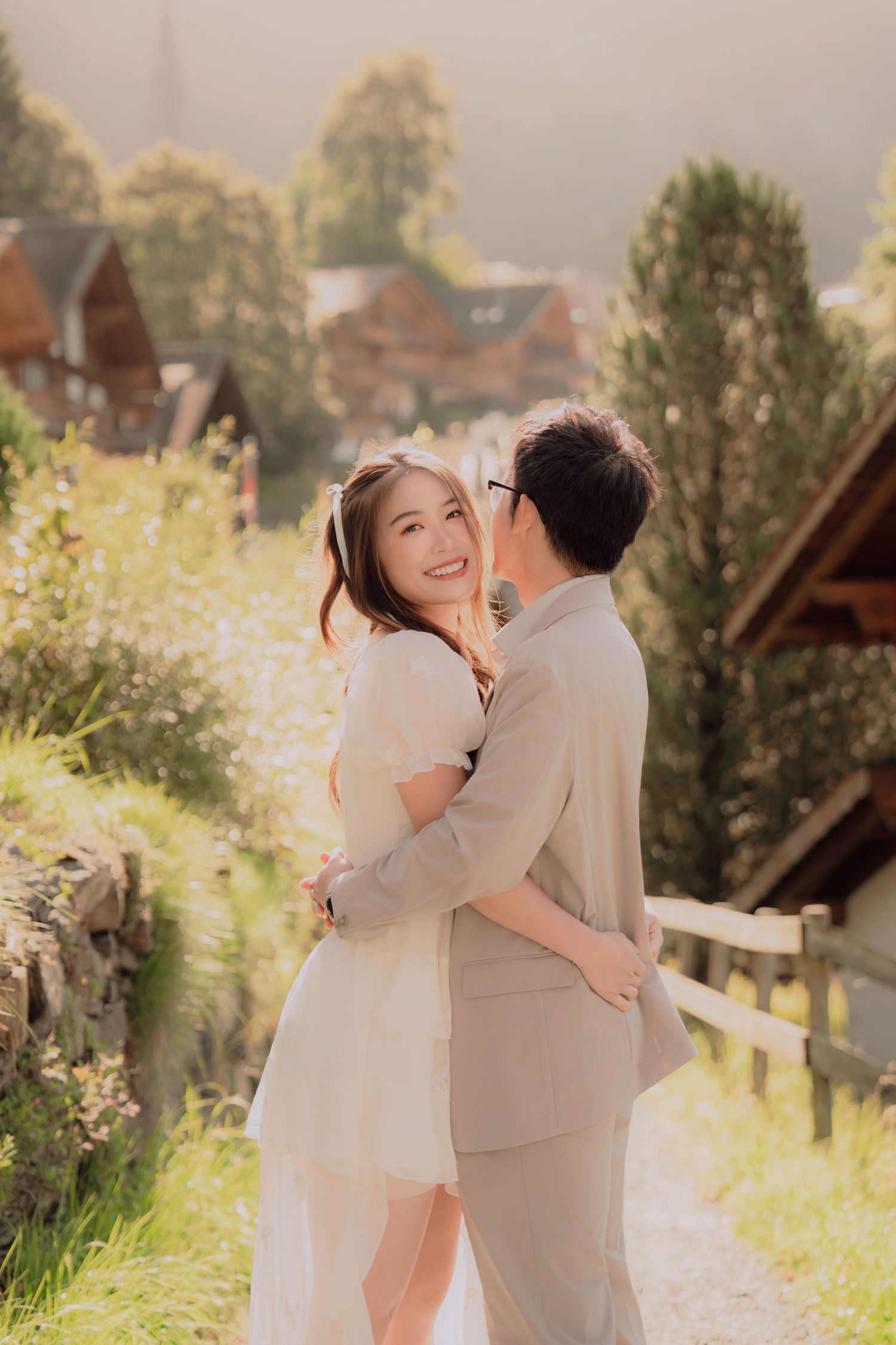 Smiling bride in white dress embracing groom on glowing sunlit path in scenic Swiss mountain village.