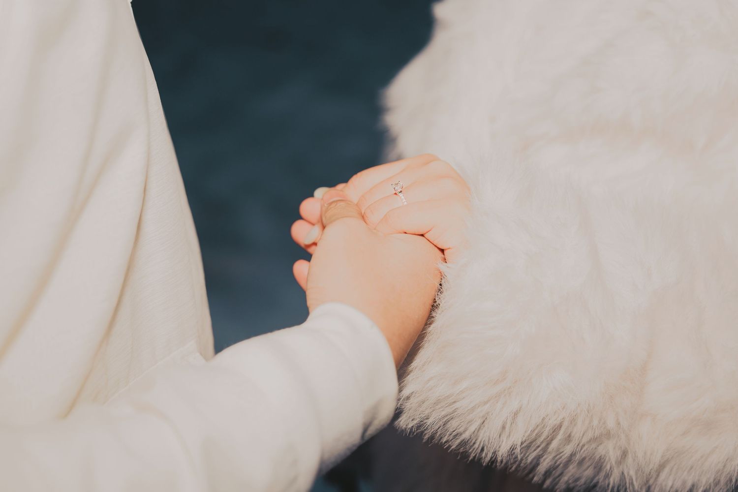 Close-up of couple holding hands wearing white outfits, with engagement ring visible on finger.