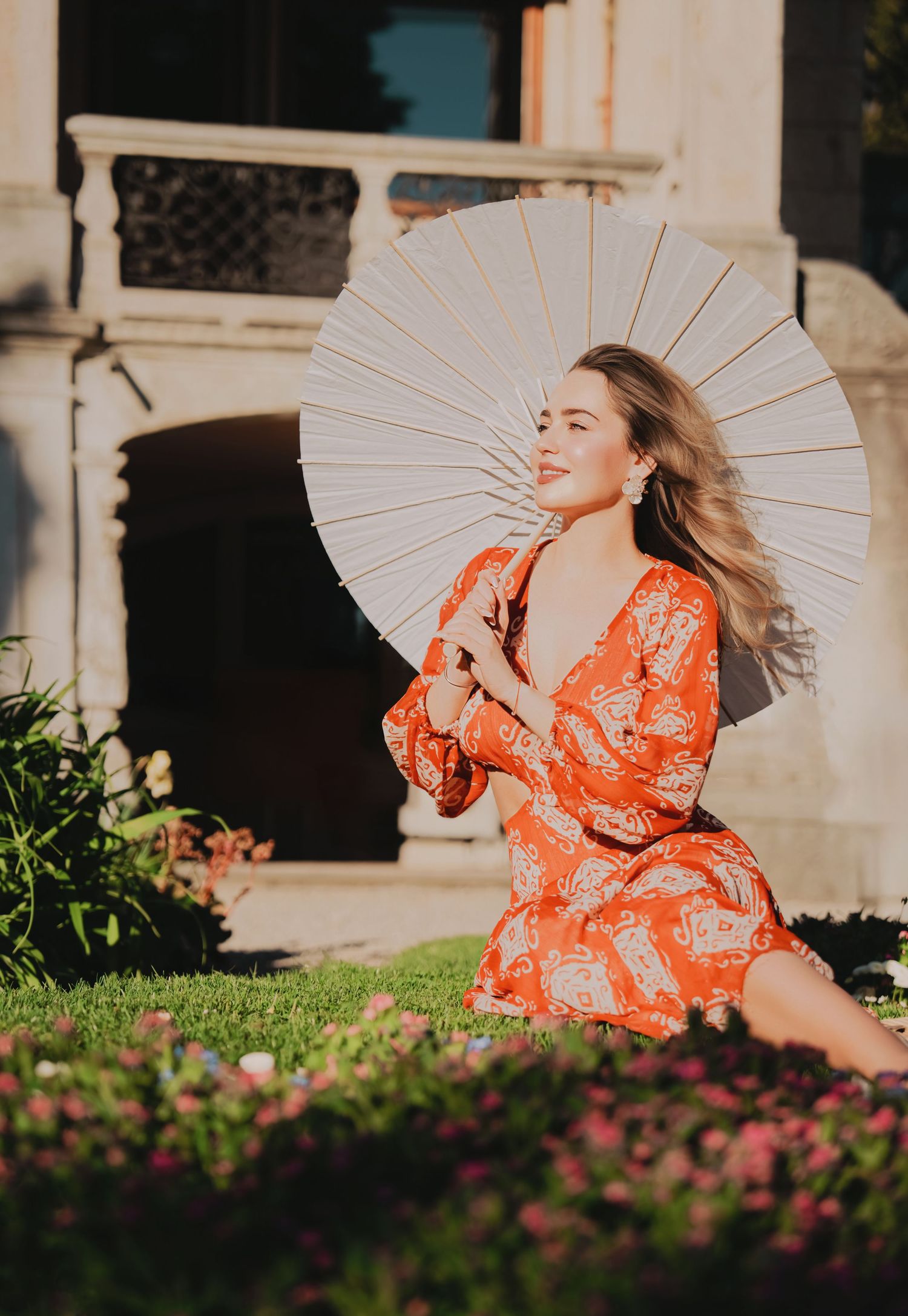 Blonde woman in orange paisley dress holding white parasol sitting on grass near historic building.
