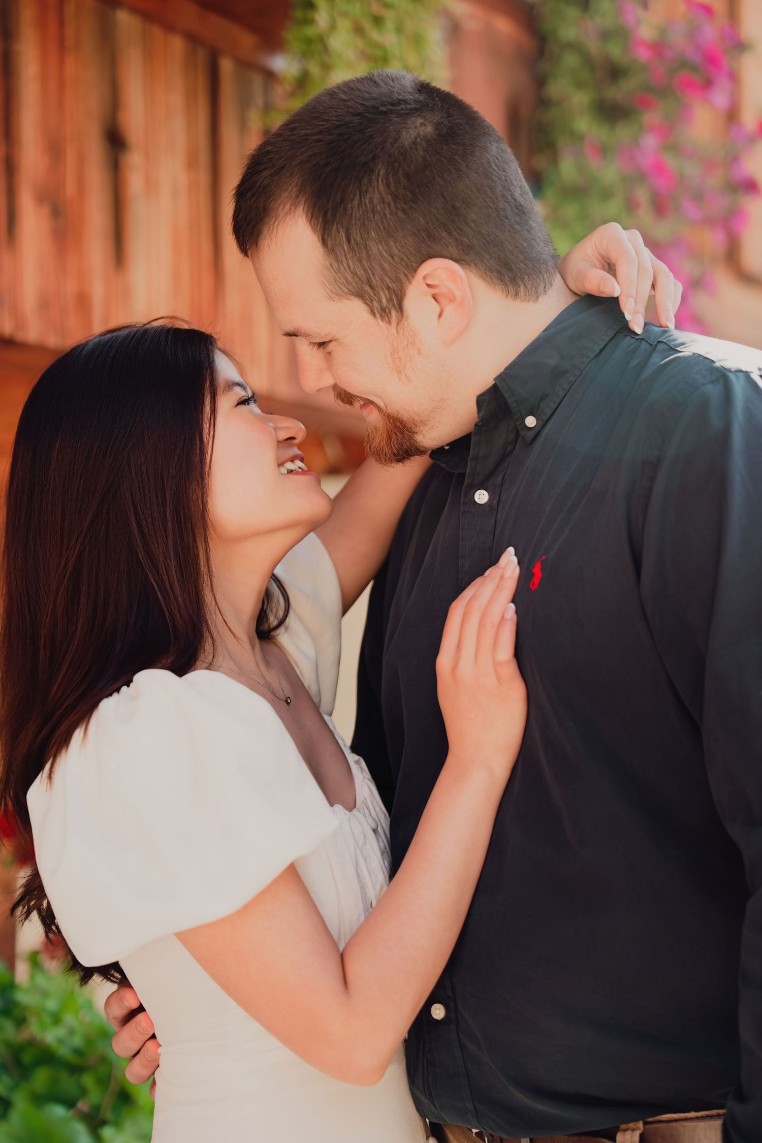 A couple almost kissing and embracing outdoors near a wooden building with colorful flowers in background.