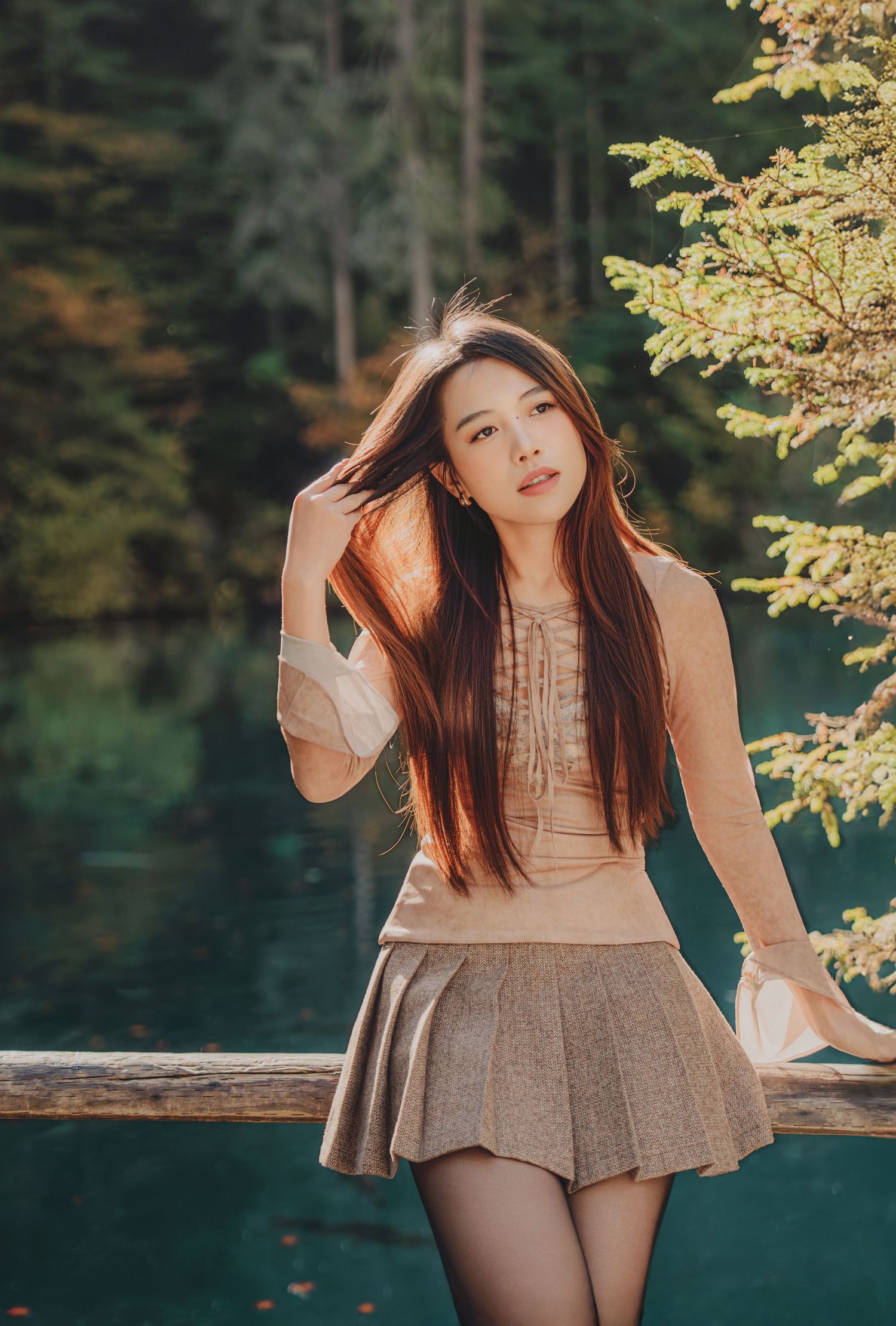 Young Asian woman in a beige top and pleated skirt posing by a wooden fence with a blue lake backdrop.