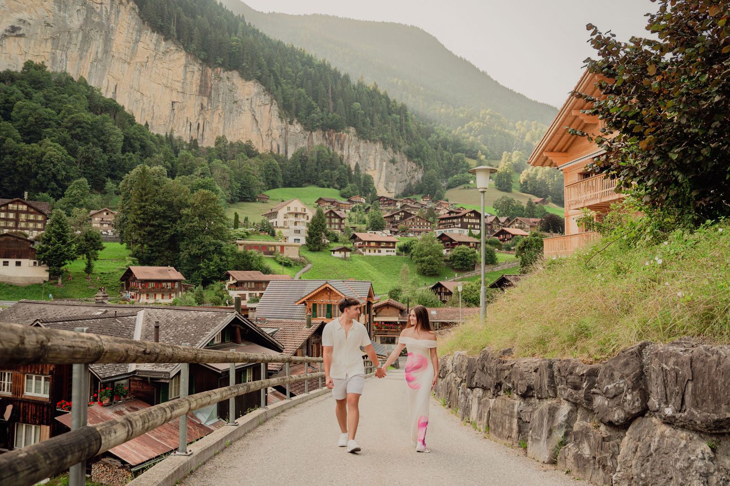Couple walking hand in hand on a bridge with a beautiful Swiss alpine village and cliffs behind them.