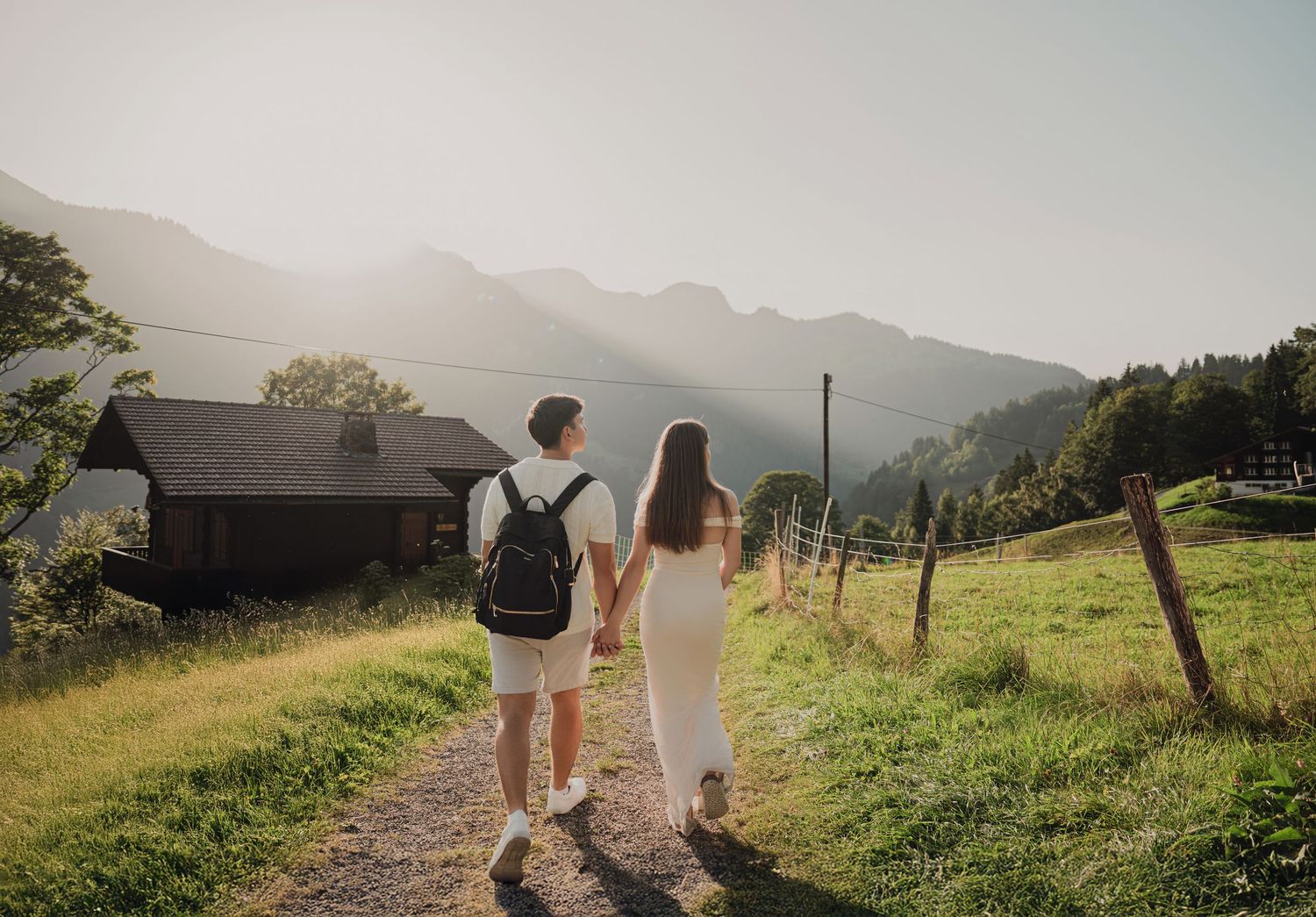 A couple walks hand in hand along a sunny mountain path toward a rustic alpine cottage.