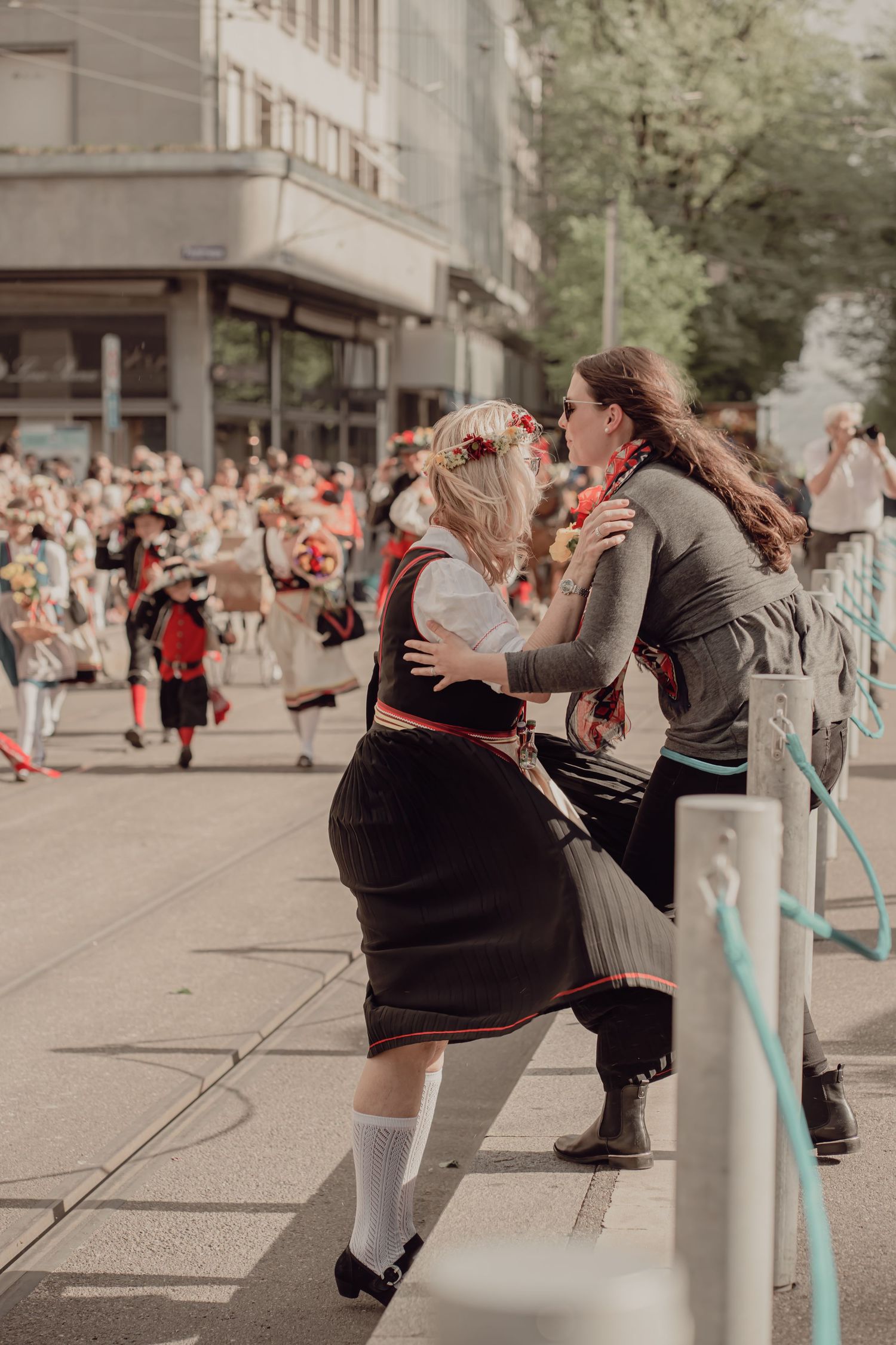 Two women in traditional European folk costumes embrace warmly during a festive street parade.