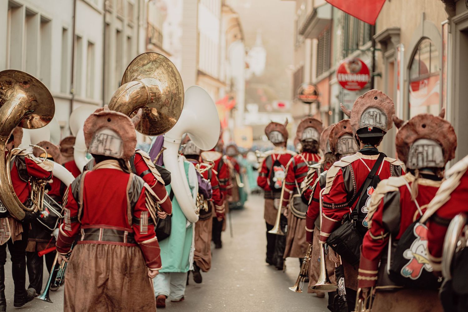 Marching band in red historical costumes parade through a European city street.