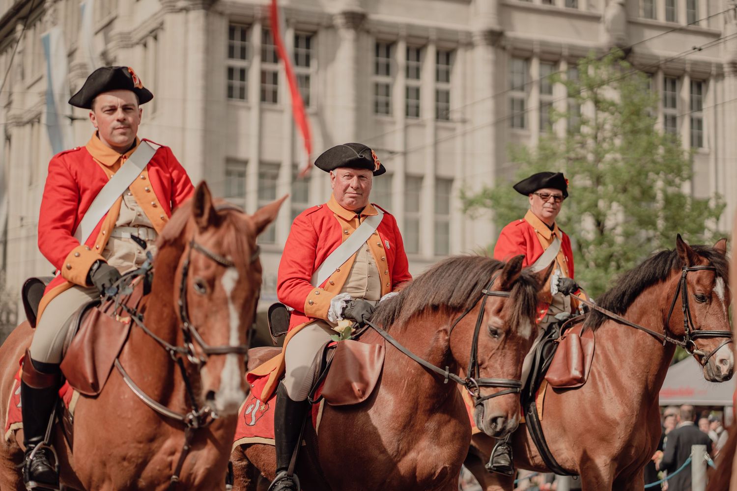 Three men in red colonial-era uniforms ride brown horses together in a historical city parade.