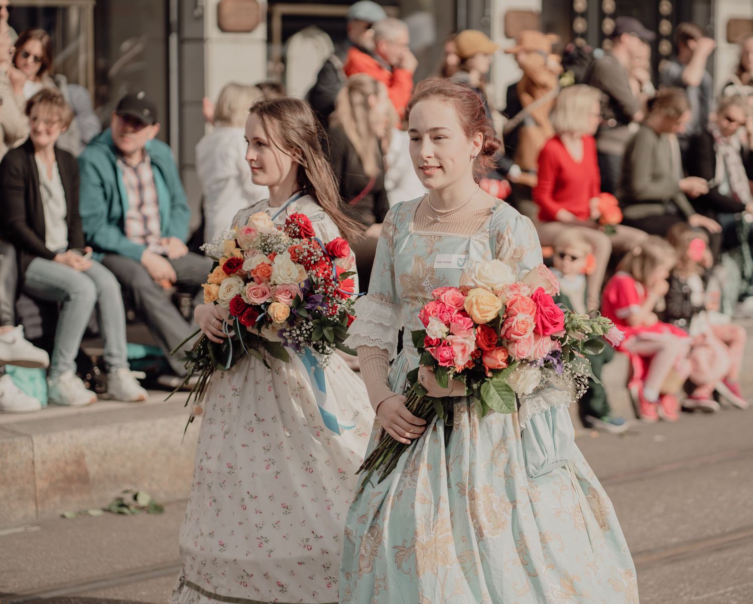 Two women in vintage floral dresses carrying colorful bouquets at a traditional street parade.